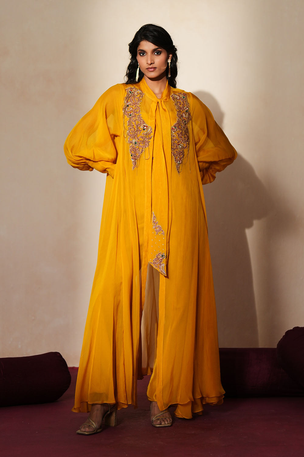 Woman in a golden yellow traditional outfit standing againsta beige wall on a red carpet.