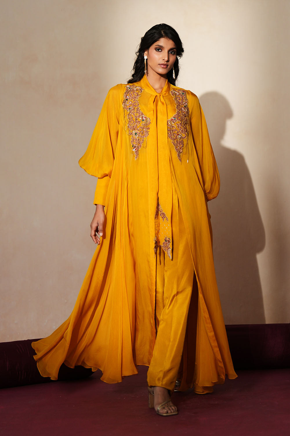 Woman in a golden yellow traditional outfit standing againsta beige wall on a red carpet.