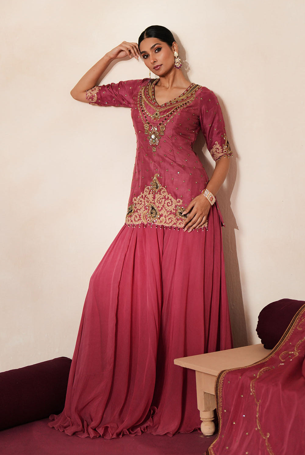 Woman in a mauve pink traditional outfit standing againsta beige wall on a red carpet.