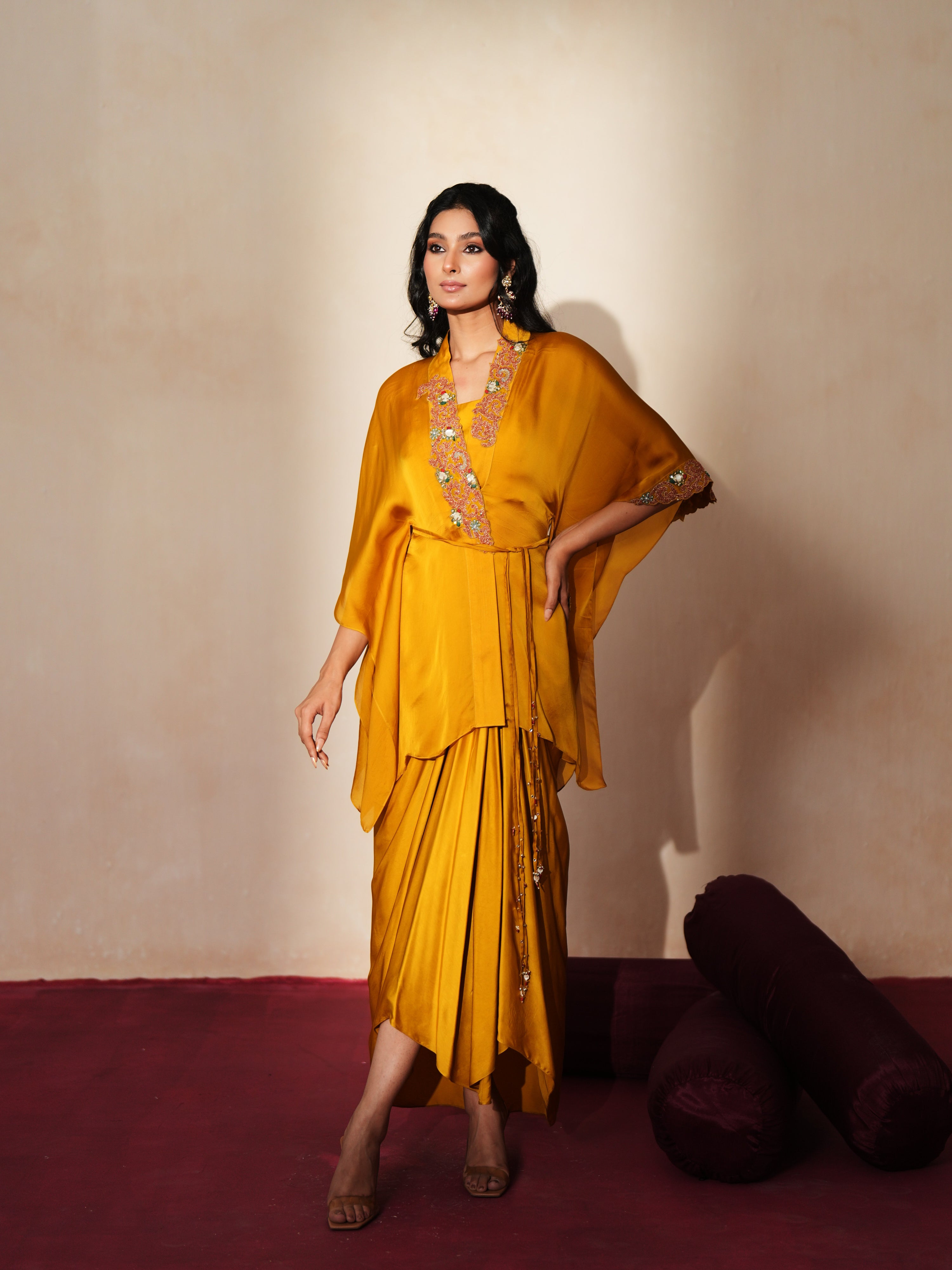 Woman in a golden yellow traditional outfit standing againsta beige wall on a red carpet.