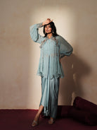 Woman in a pastel blue traditional outfit standing againsta beige wall on a red carpet.