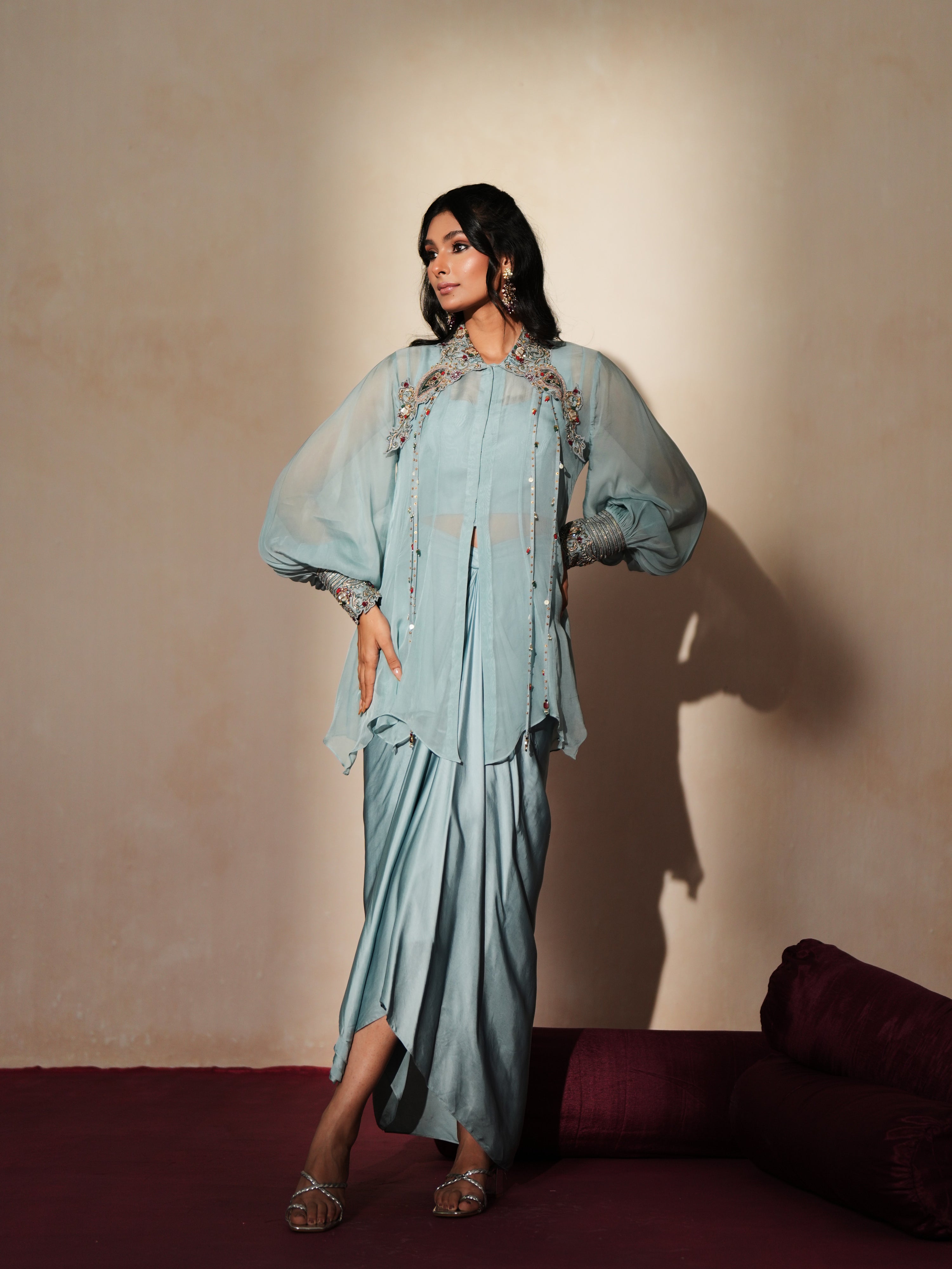 Woman in a pastel blue traditional outfit standing againsta beige wall on a red carpet.