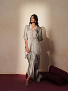 Woman in a mettalic grey traditional outfit standing againsta beige wall on a red carpet.