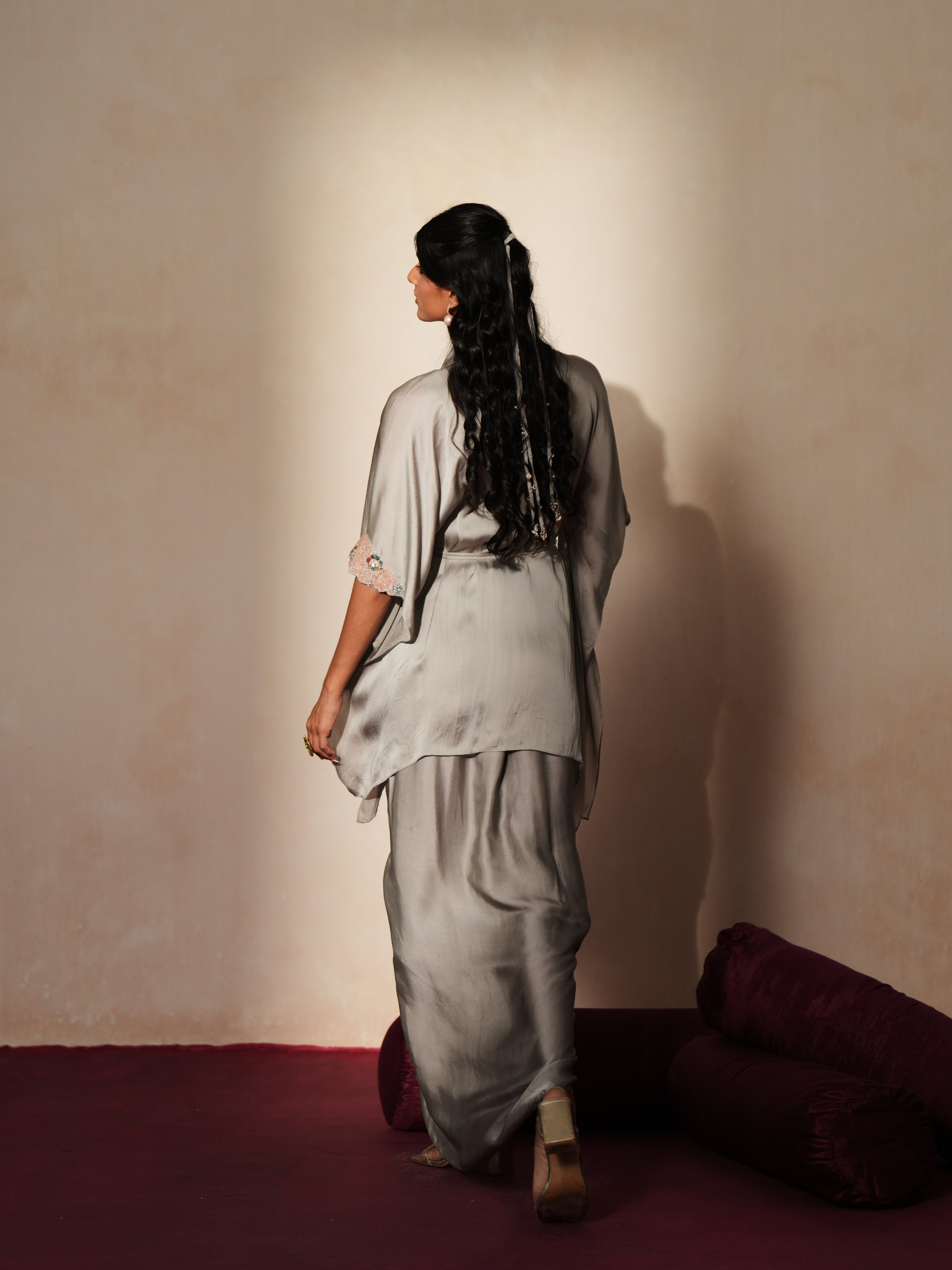 Woman in a mettalic grey traditional outfit standing againsta beige wall on a red carpet.