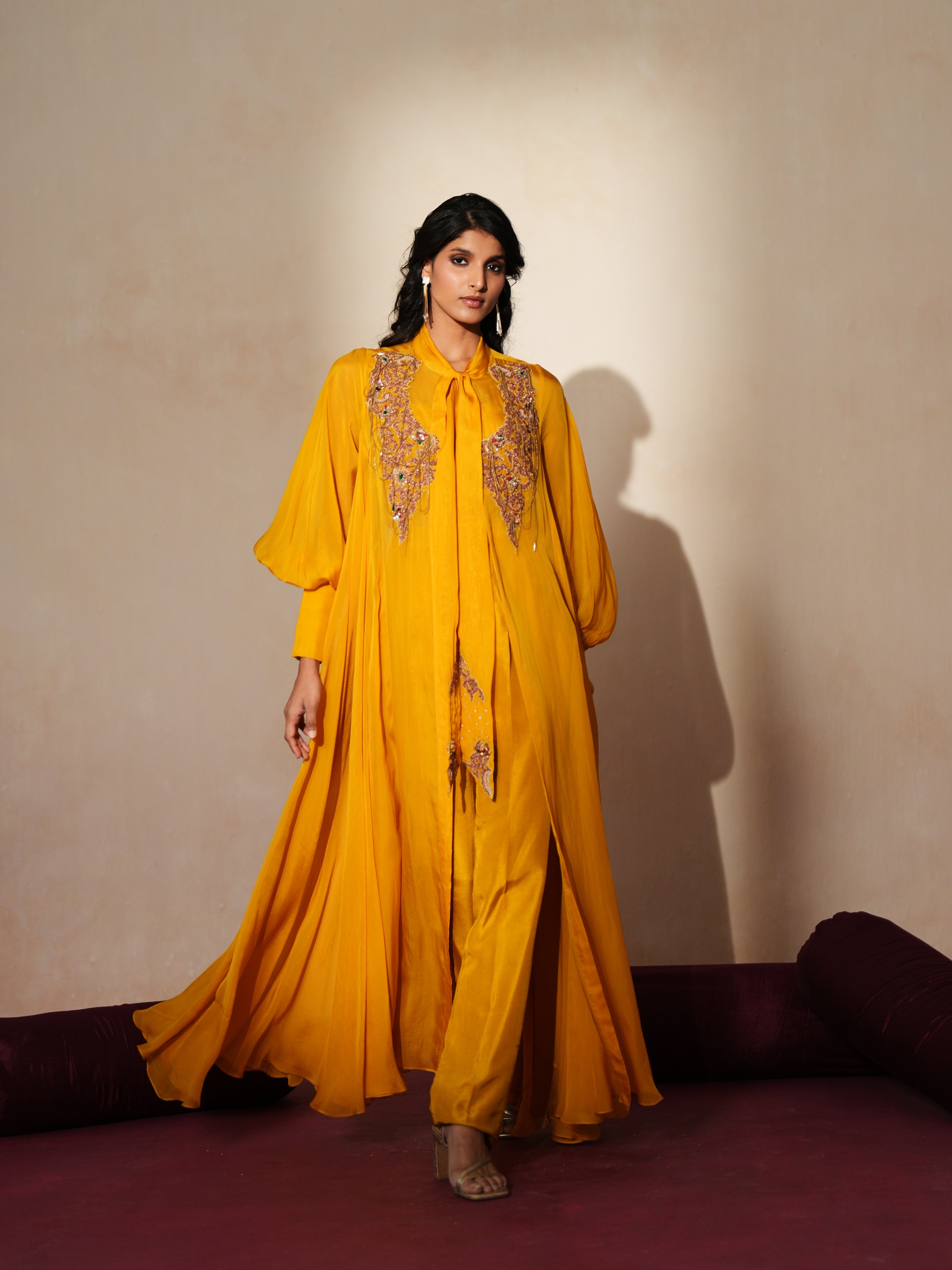 Woman in a golden yellow traditional outfit standing againsta beige wall on a red carpet.