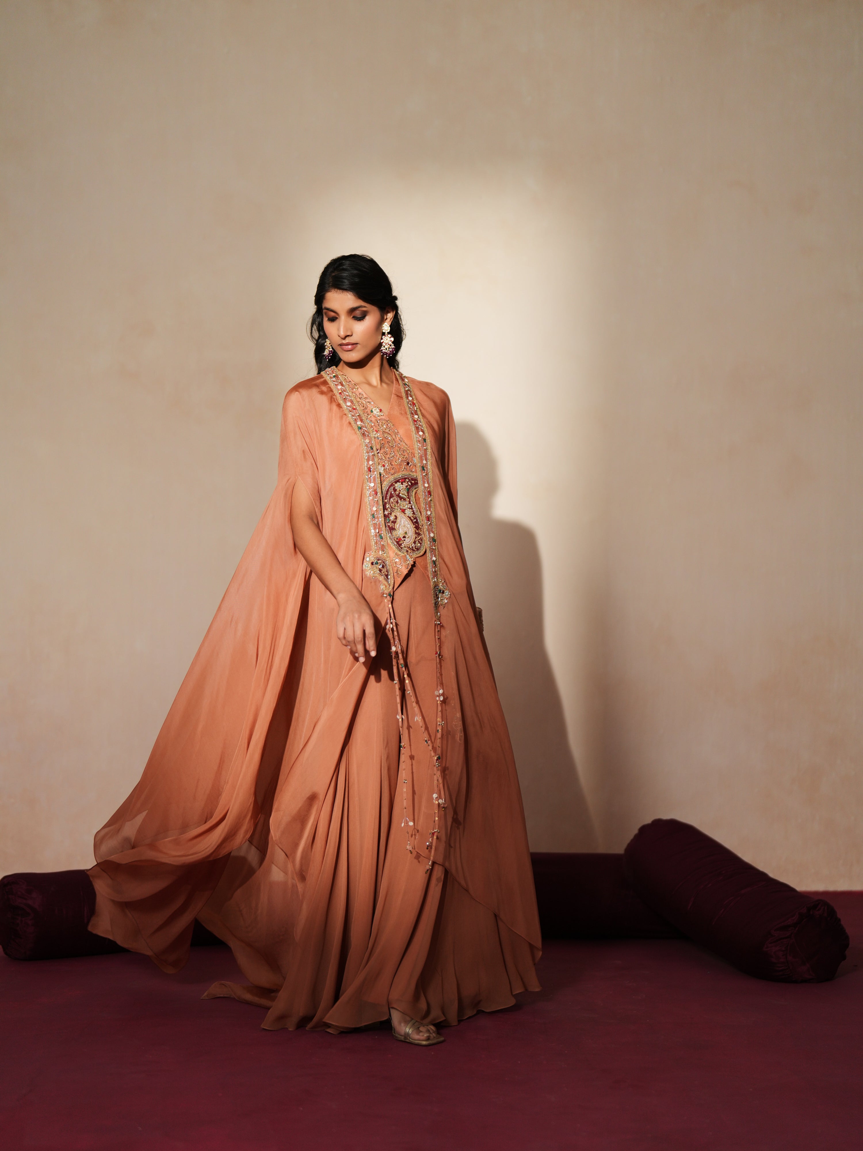 Woman in a metallic mocha  traditional outfit standing againsta beige wall on a red carpet.