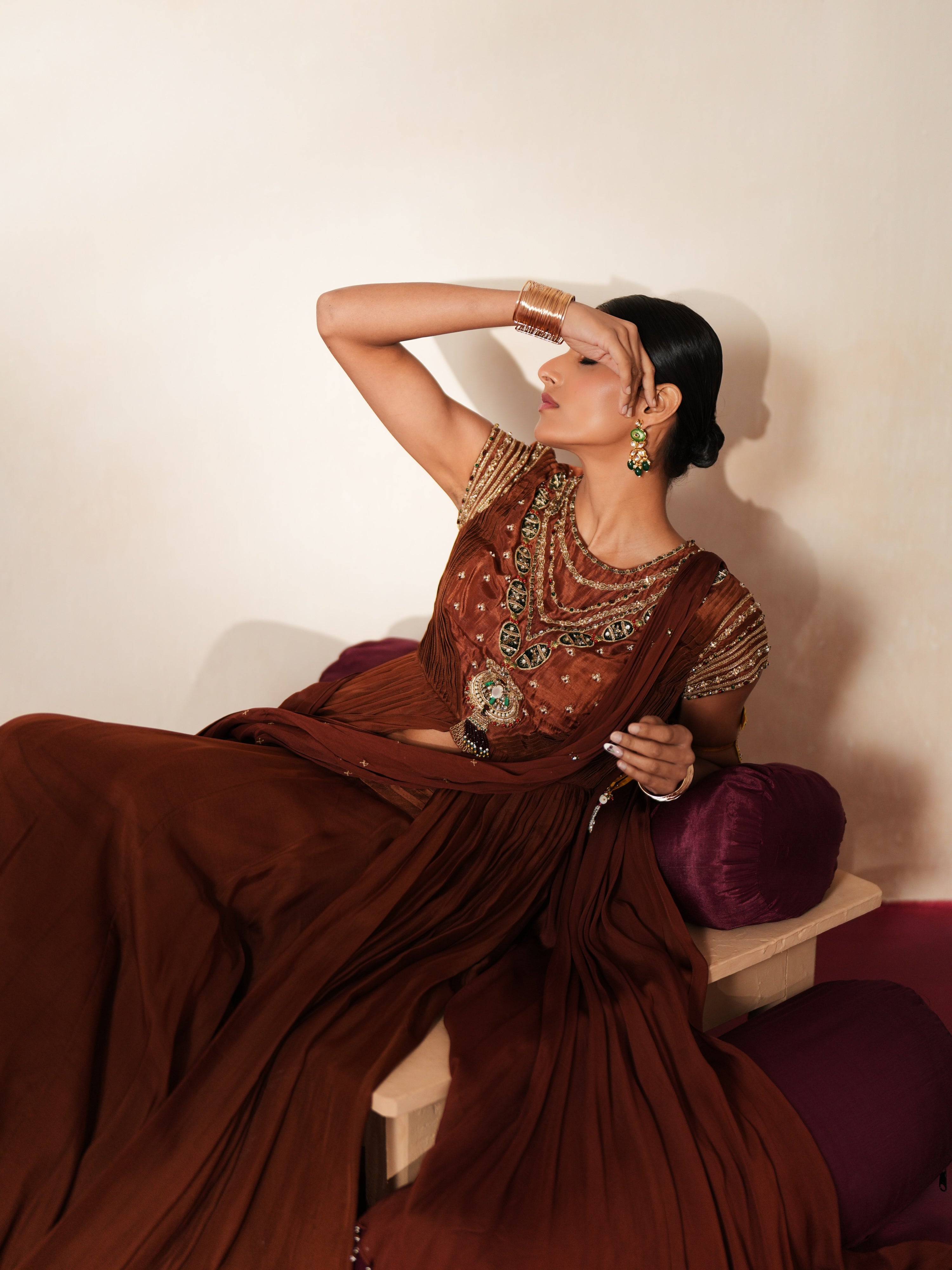 Woman in a mettalic brown traditional outfit standing againsta beige wall on a red carpet.