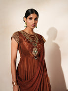 Woman in a mettalic brown traditional outfit standing againsta beige wall on a red carpet.