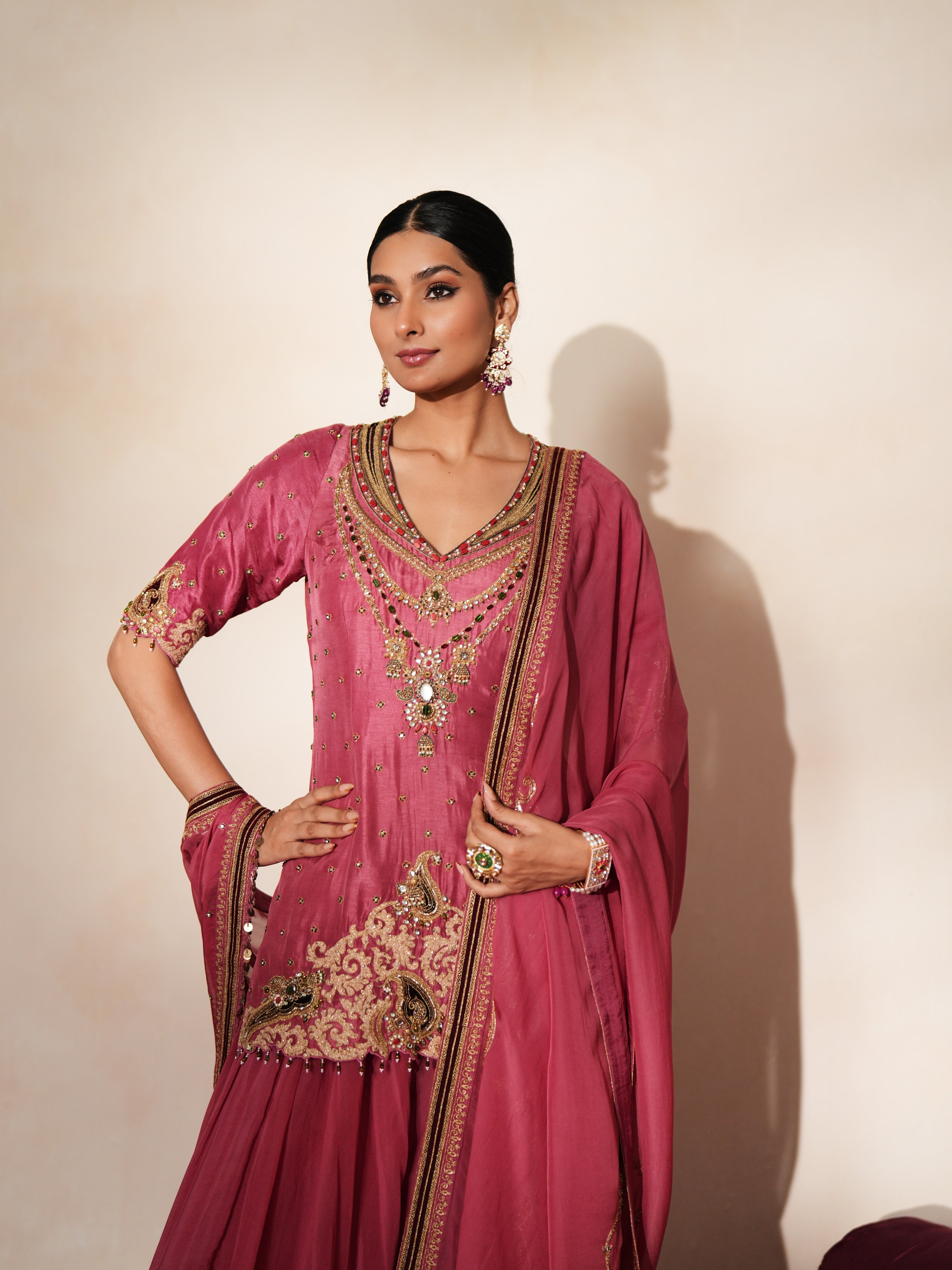 Woman in a mauve pink traditional outfit standing againsta beige wall on a red carpet.