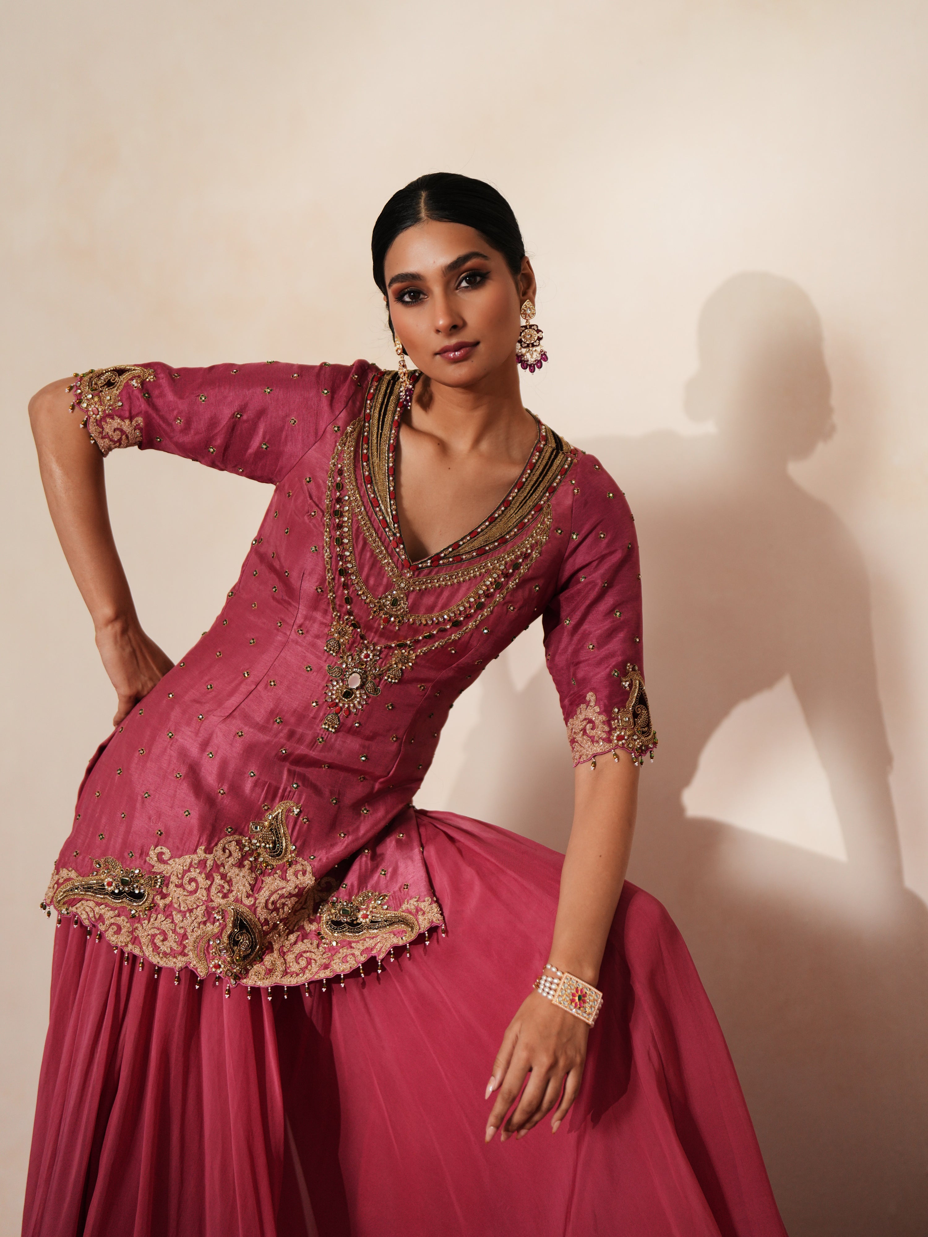 Woman in a mauve pink traditional outfit standing againsta beige wall on a red carpet.