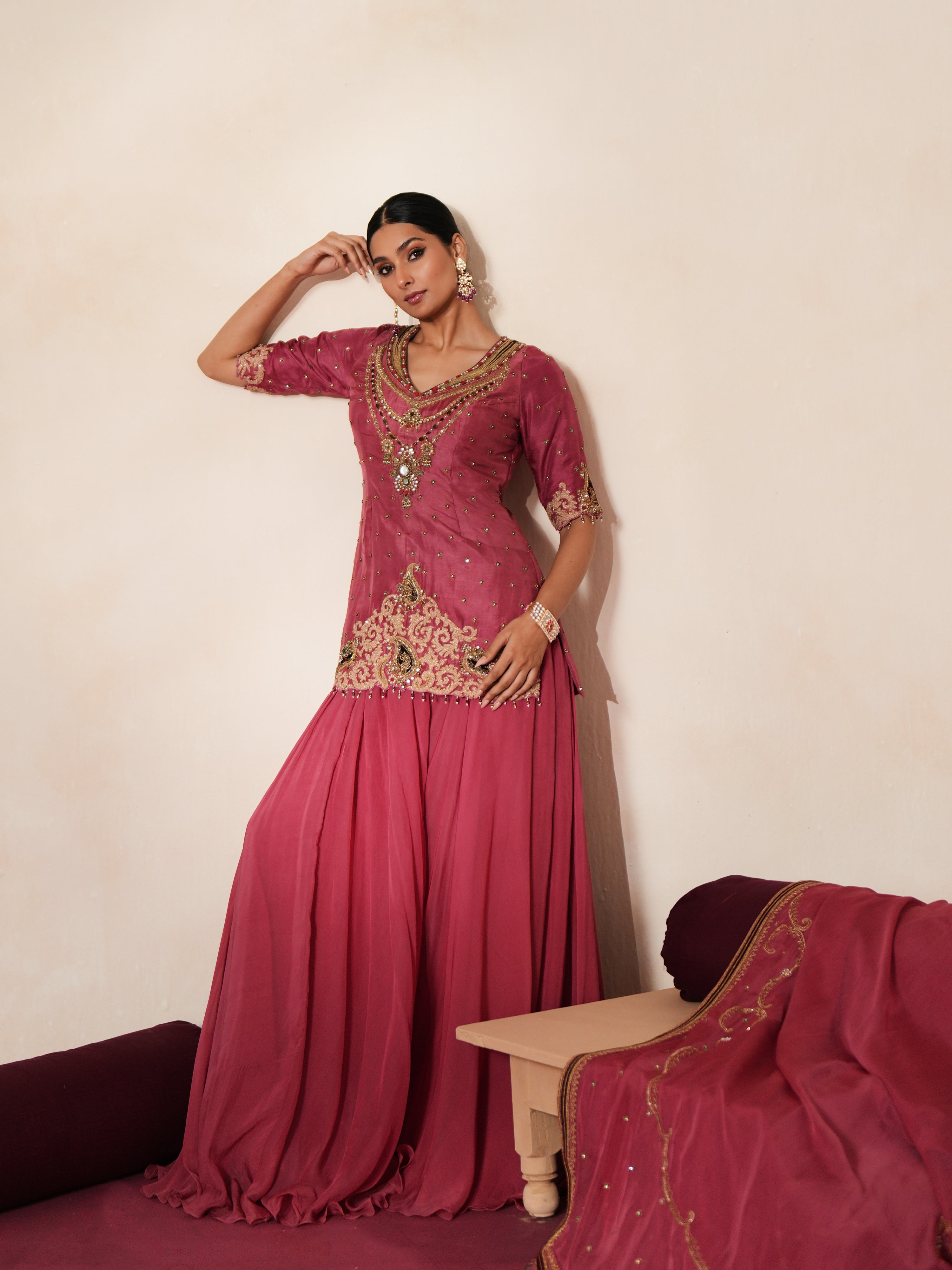 Woman in a mauve pink traditional outfit standing againsta beige wall on a red carpet.