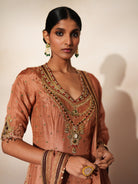 Woman in a metallic mocha  traditional outfit standing againsta beige wall on a red carpet.