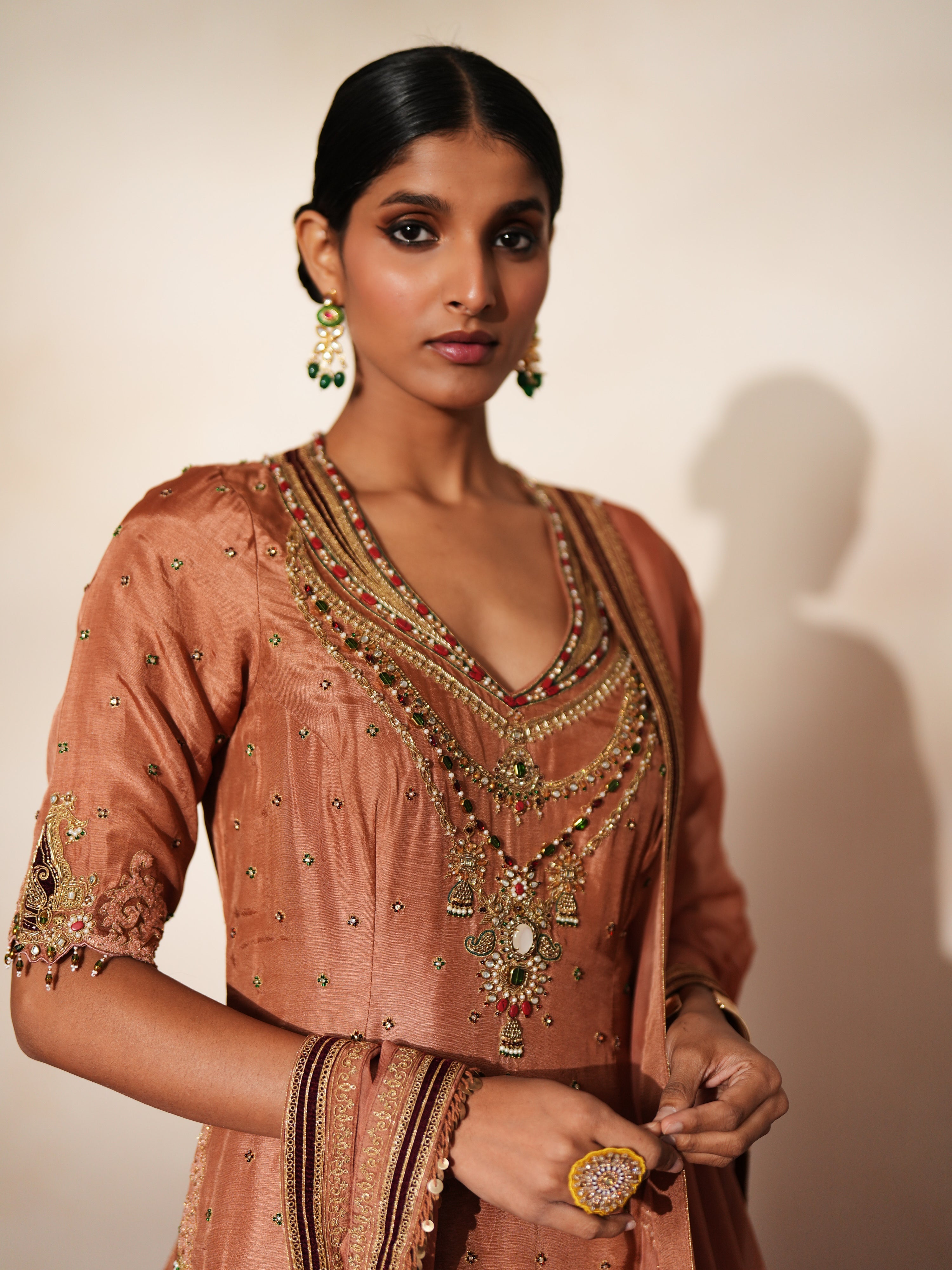 Woman in a metallic mocha  traditional outfit standing againsta beige wall on a red carpet.