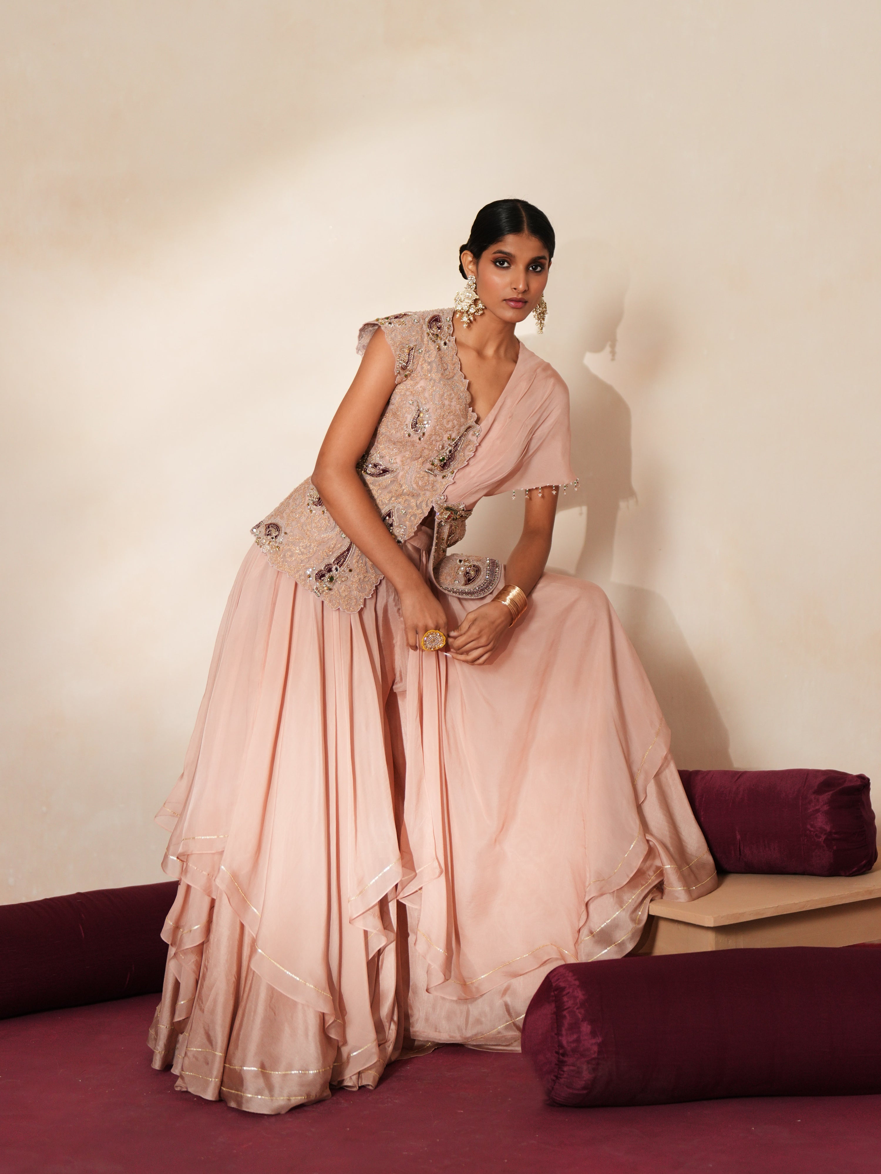 Woman in a soft pink traditional outfit standing againsta beige wall on a red carpet.