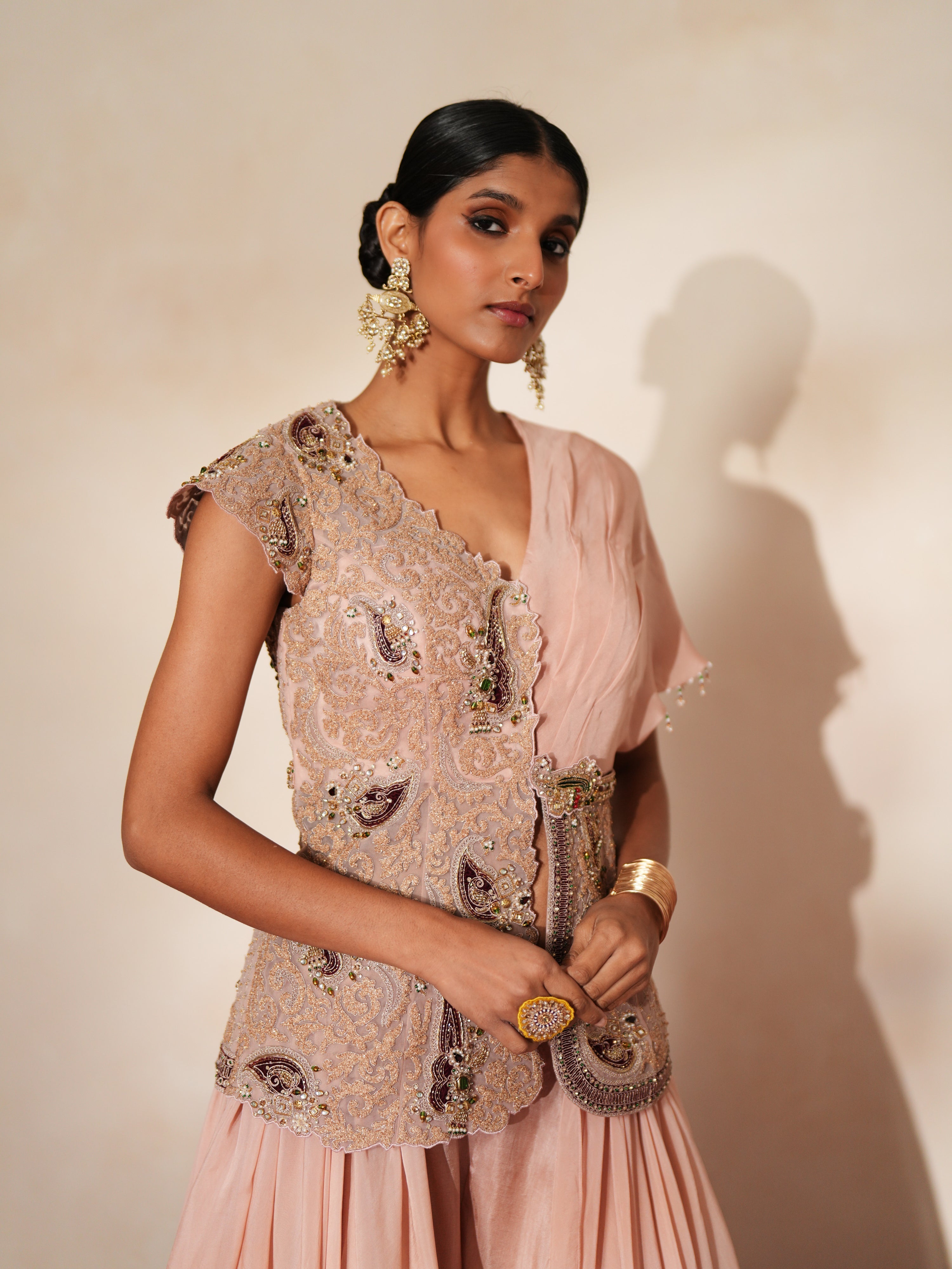 Woman in a soft pink traditional outfit standing againsta beige wall on a red carpet.
