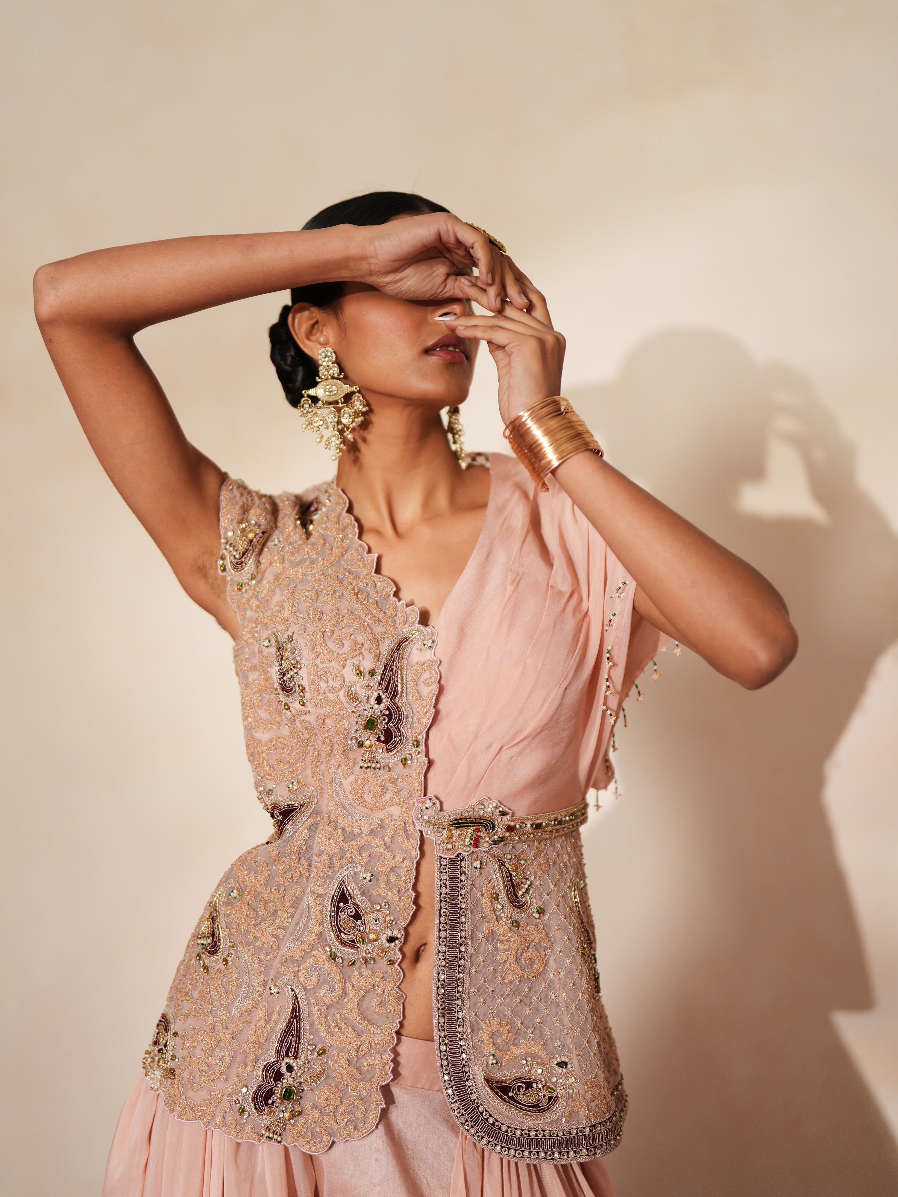Woman in a soft pink traditional outfit standing againsta beige wall on a red carpet.