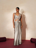 Woman in a mettalic grey traditional outfit standing againsta beige wall on a red carpet.