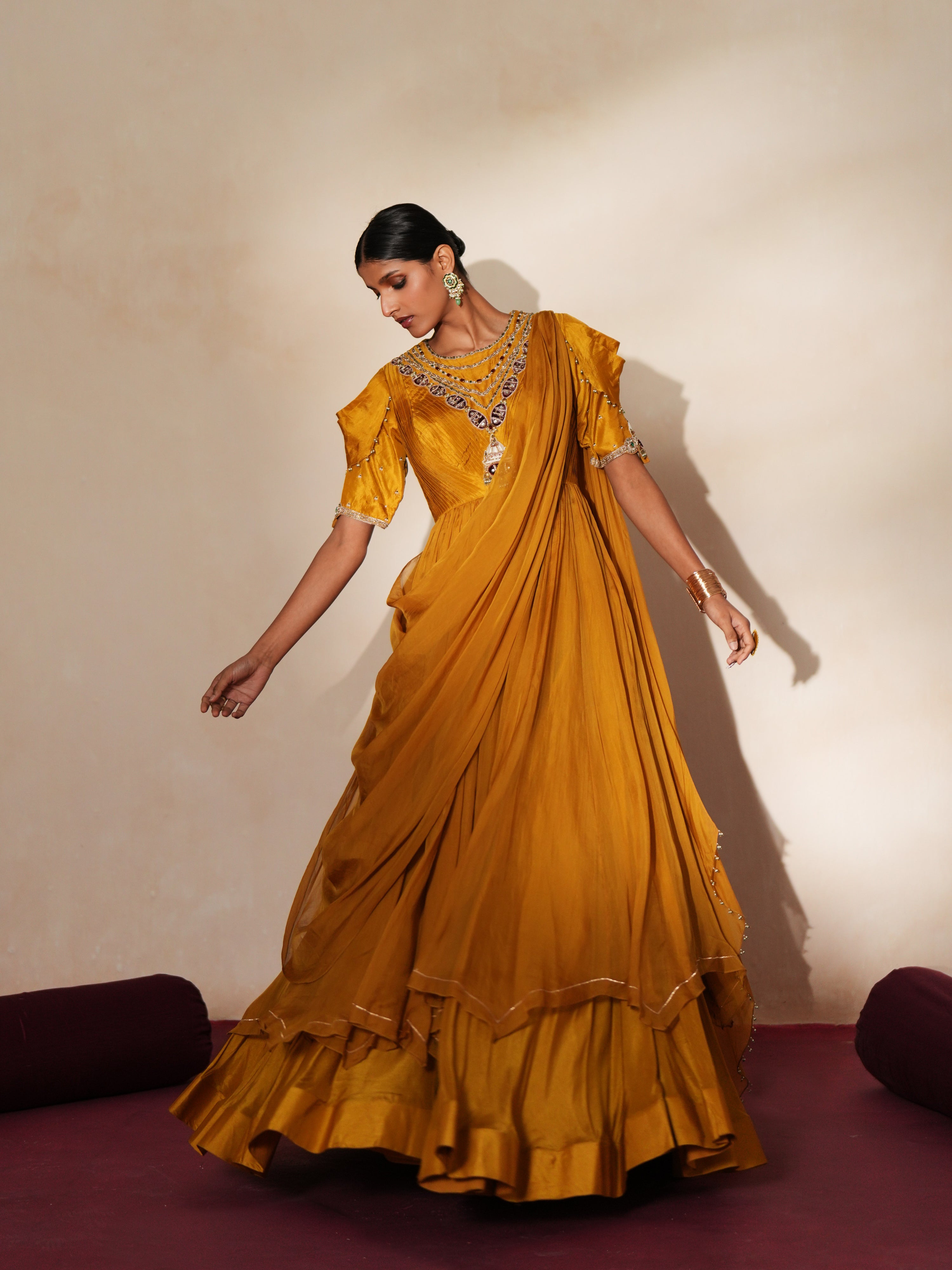 Woman in a golden yellow traditional outfit standing againsta beige wall on a red carpet.