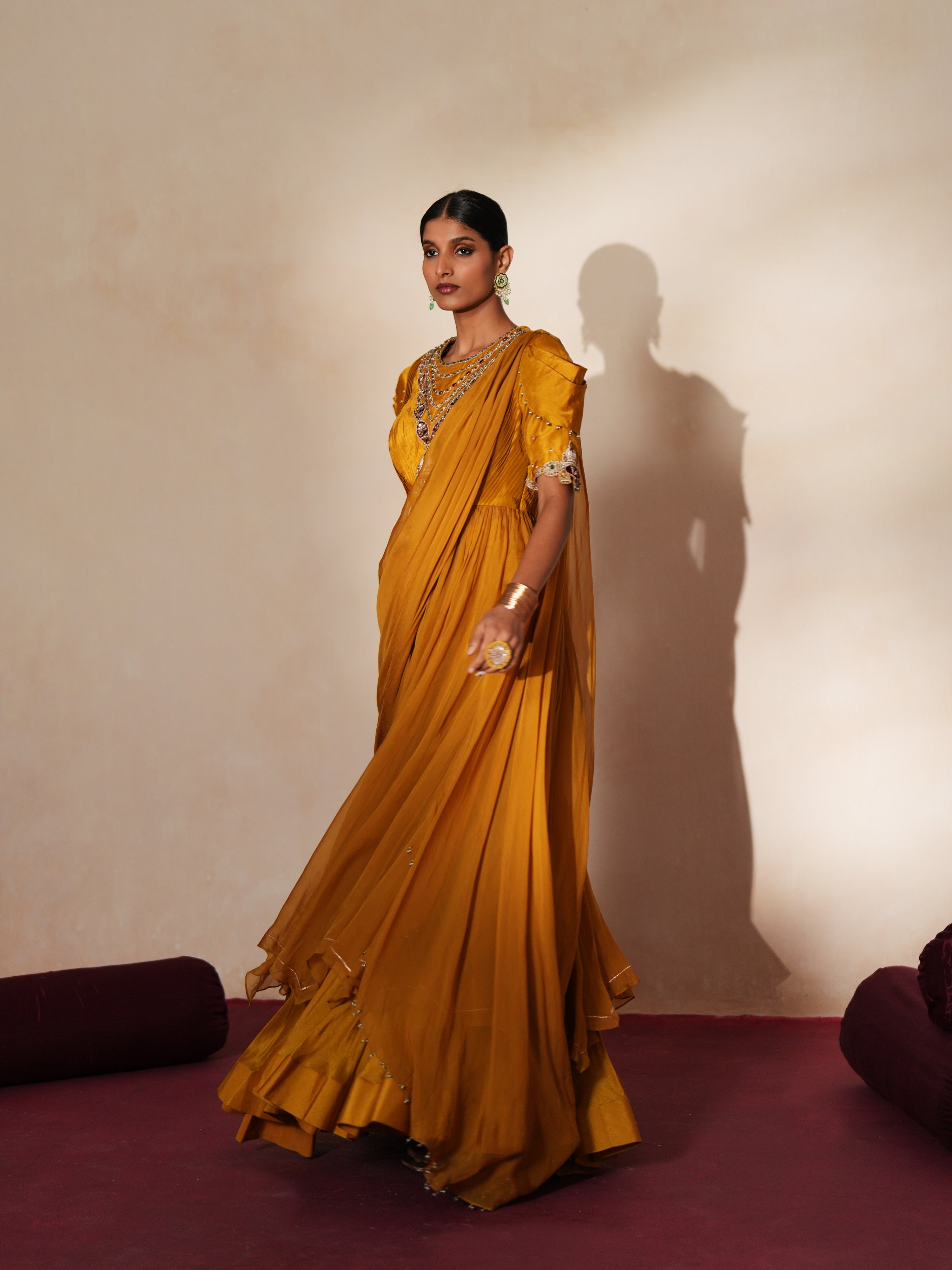 Woman in a golden yellow traditional outfit standing againsta beige wall on a red carpet.