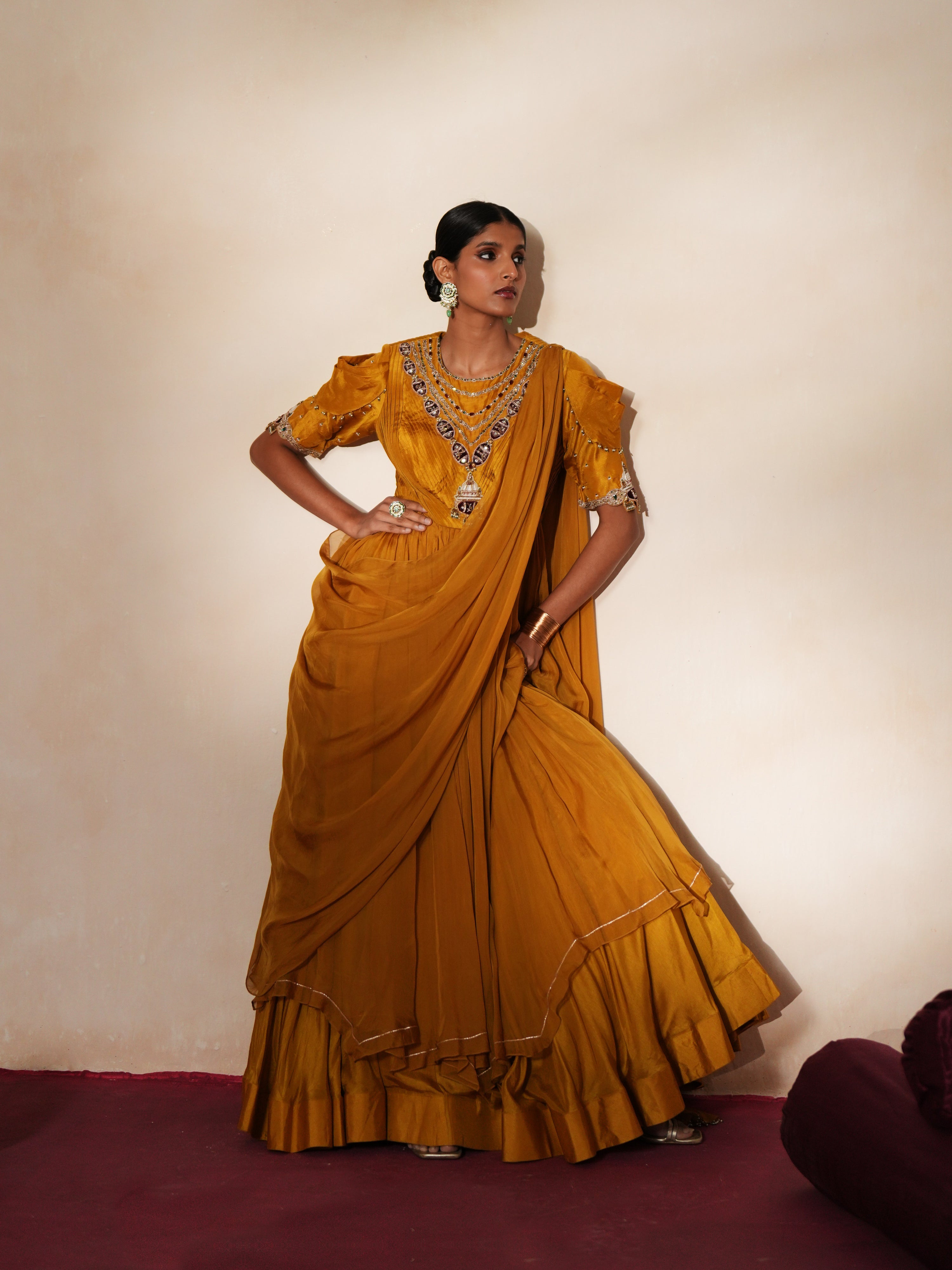 Woman in a golden yellow traditional outfit standing againsta beige wall on a red carpet.
