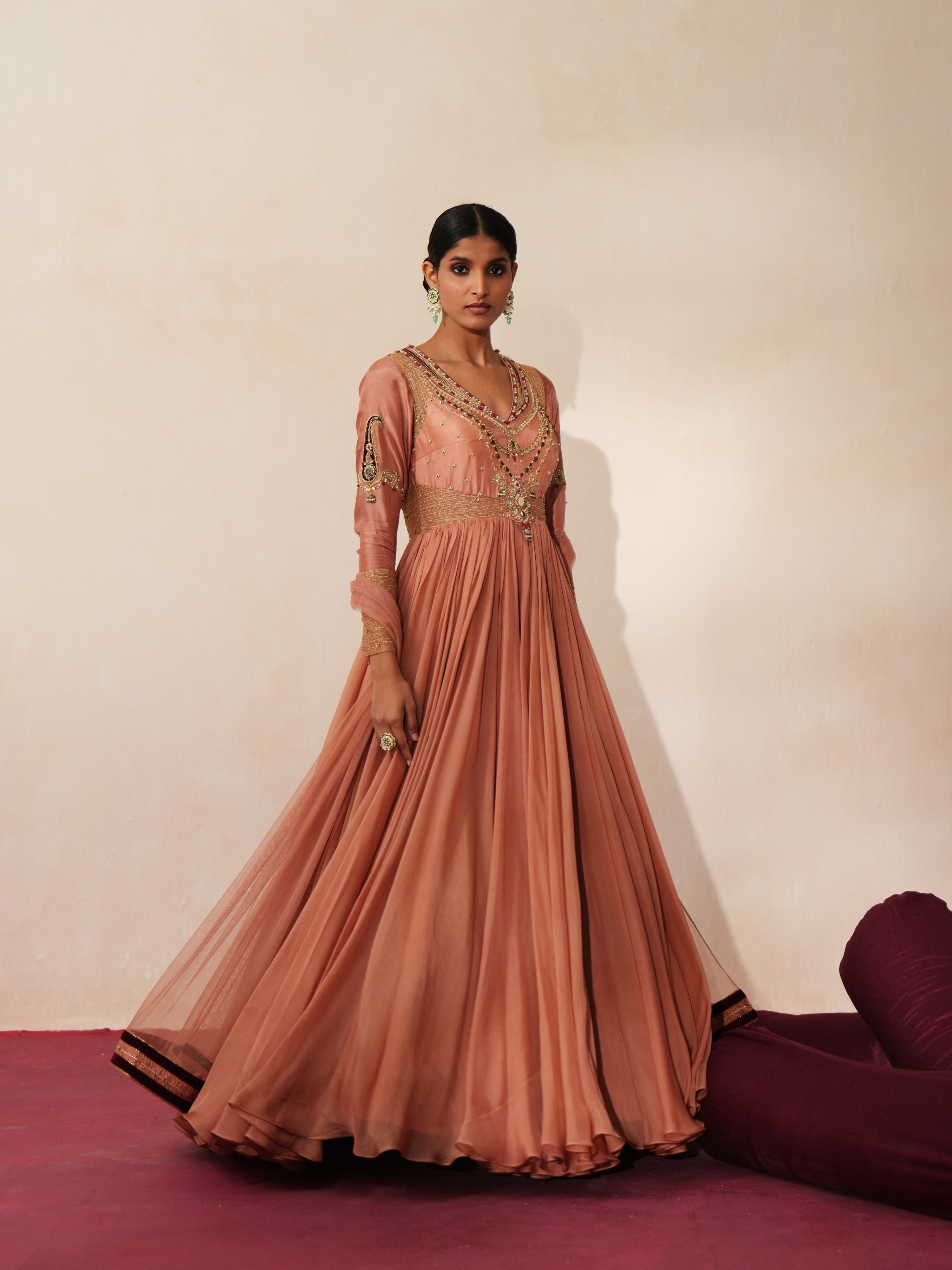 Woman in a metallic mocha  traditional outfit standing againsta beige wall on a red carpet.