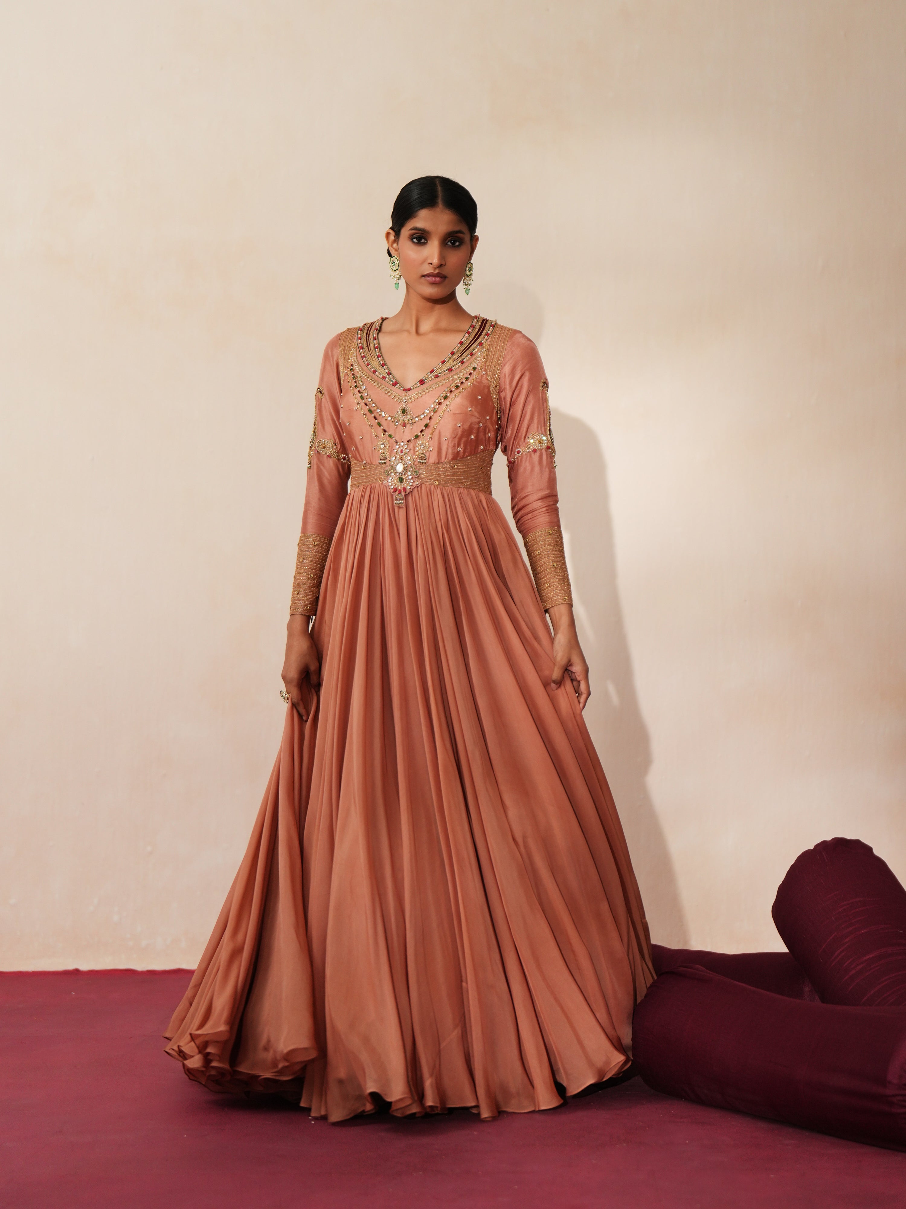 Woman in a metallic mocha  traditional outfit standing againsta beige wall on a red carpet.