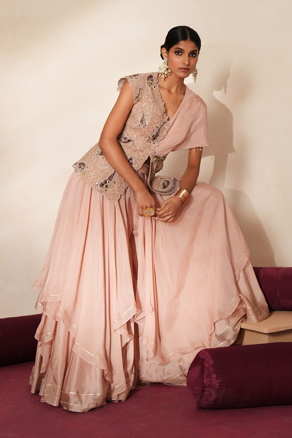 Woman in a soft pink traditional outfit standing againsta beige wall on a red carpet.