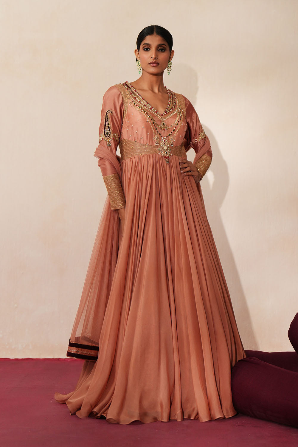 Woman in a metallic mocha  traditional outfit standing againsta beige wall on a red carpet.