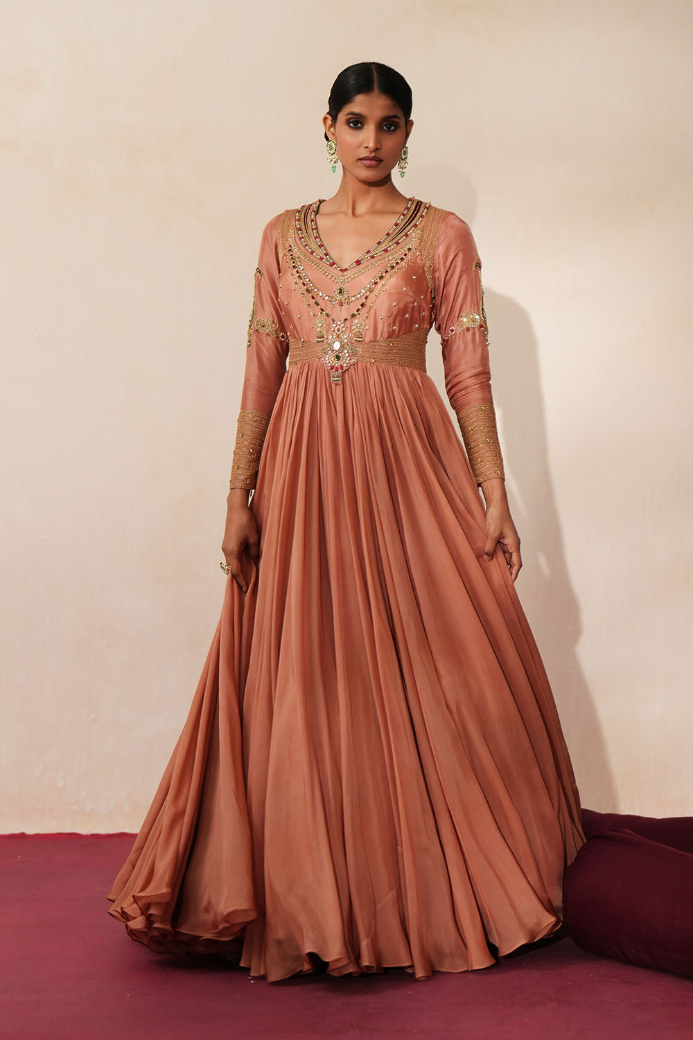 Woman in a metallic mocha  traditional outfit standing againsta beige wall on a red carpet.