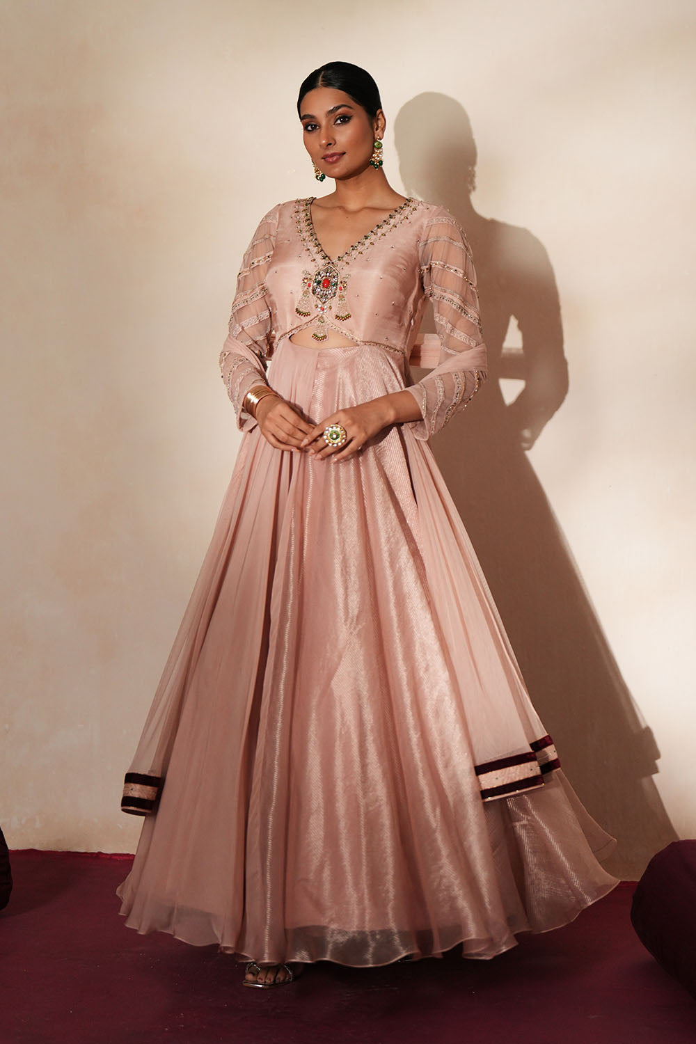 Woman in a soft pink traditional outfit standing againsta beige wall on a red carpet.