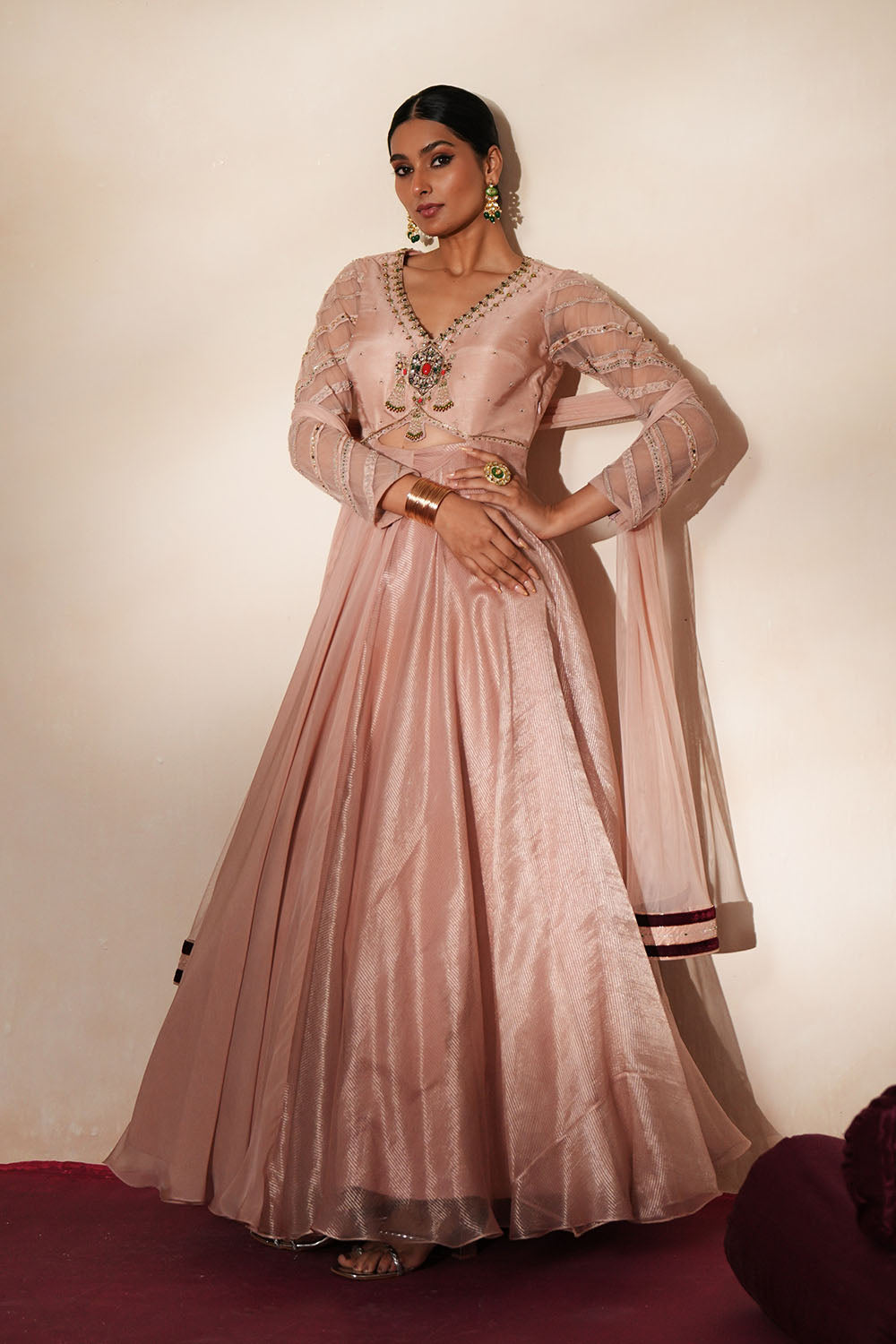 Woman in a soft pink traditional outfit standing againsta beige wall on a red carpet.