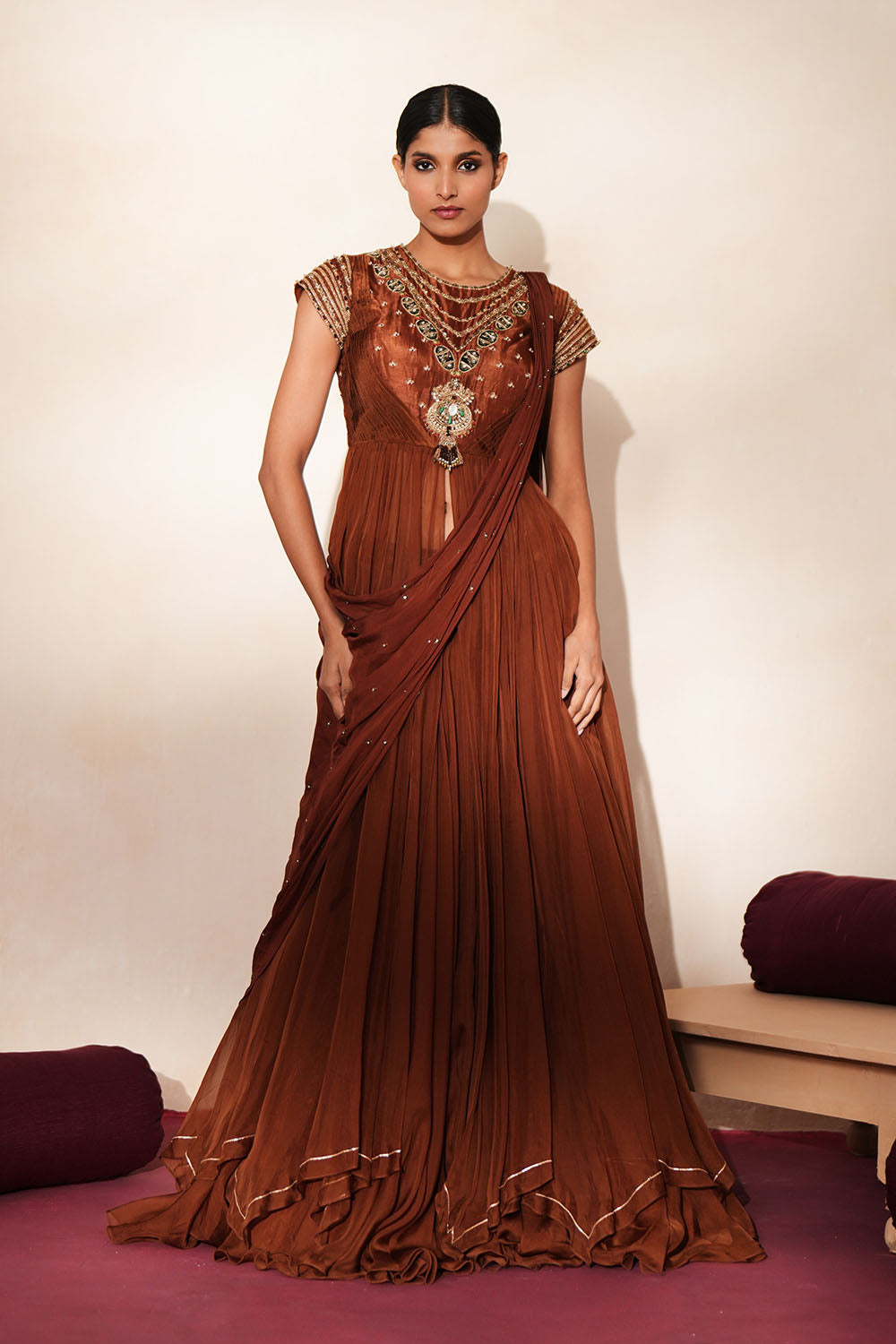 Woman in a mettalic brown traditional outfit standing againsta beige wall on a red carpet.