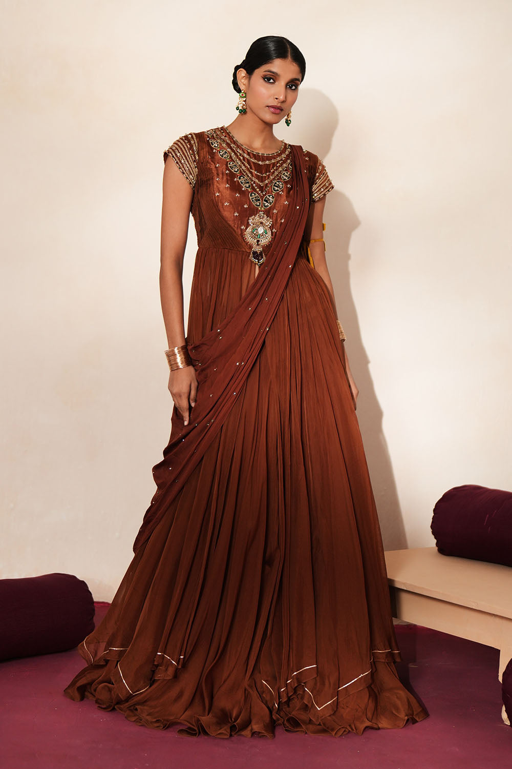 Woman in a mettalic brown traditional outfit standing againsta beige wall on a red carpet.