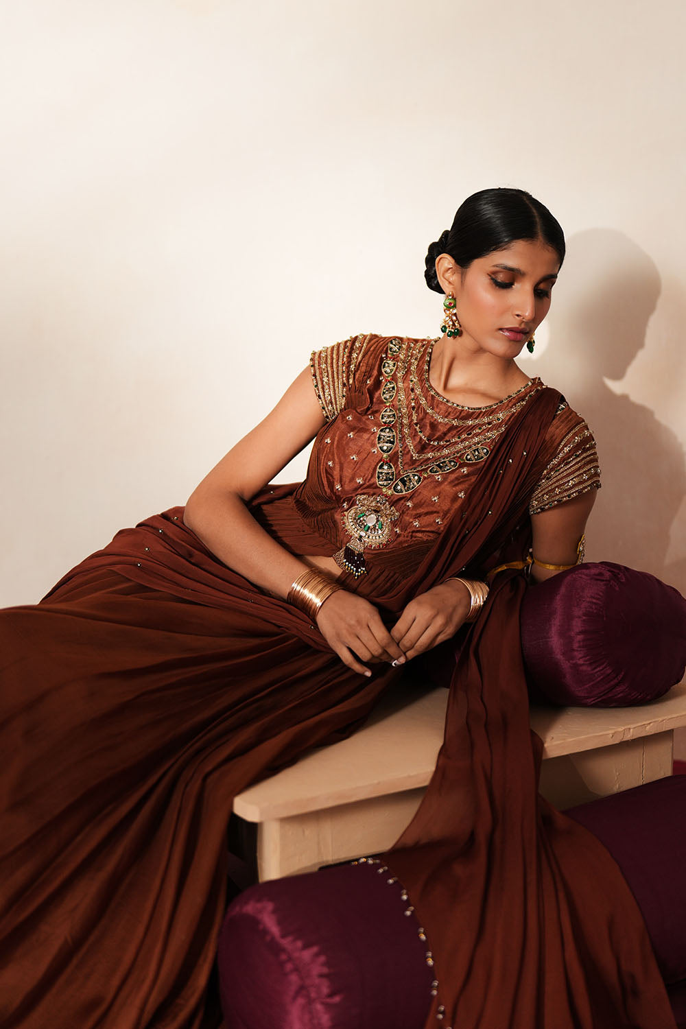 Woman in a mettalic brown traditional outfit standing againsta beige wall on a red carpet.