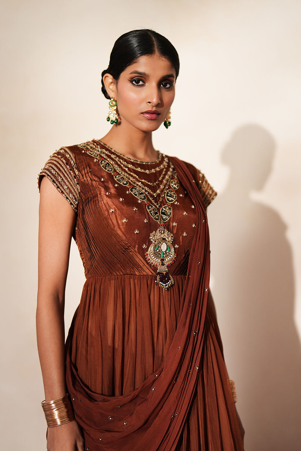 Woman in a mettalic brown traditional outfit standing againsta beige wall on a red carpet.