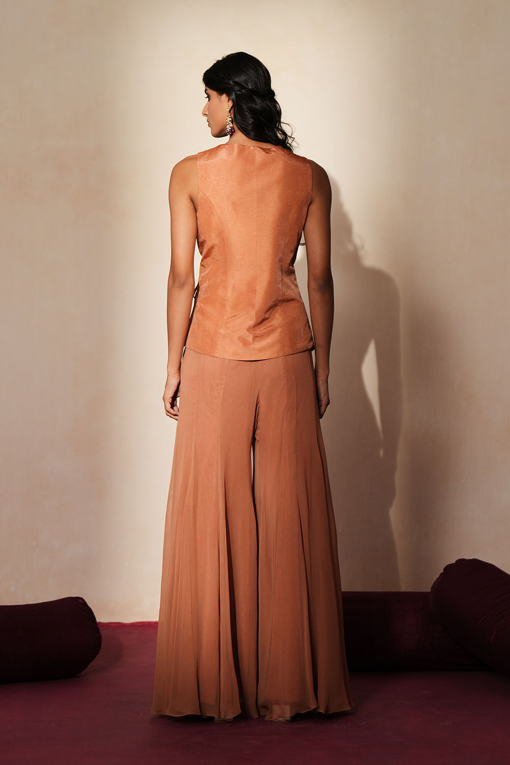 Woman in a metallic mocha  traditional outfit standing againsta beige wall on a red carpet.