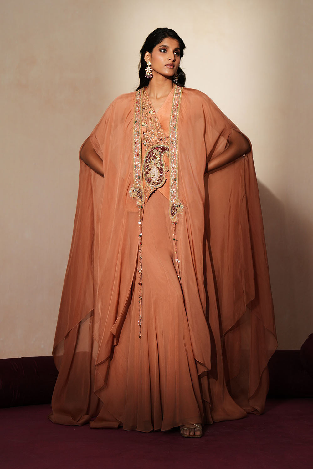 Woman in a metallic mocha  traditional outfit standing againsta beige wall on a red carpet.