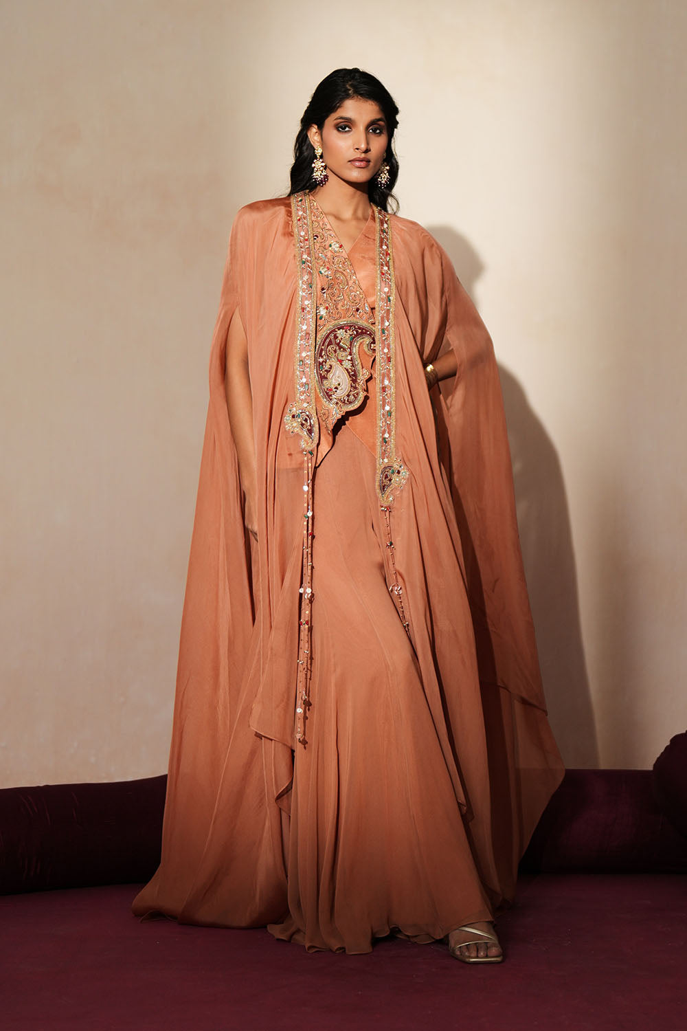 Woman in a metallic mocha  traditional outfit standing againsta beige wall on a red carpet.