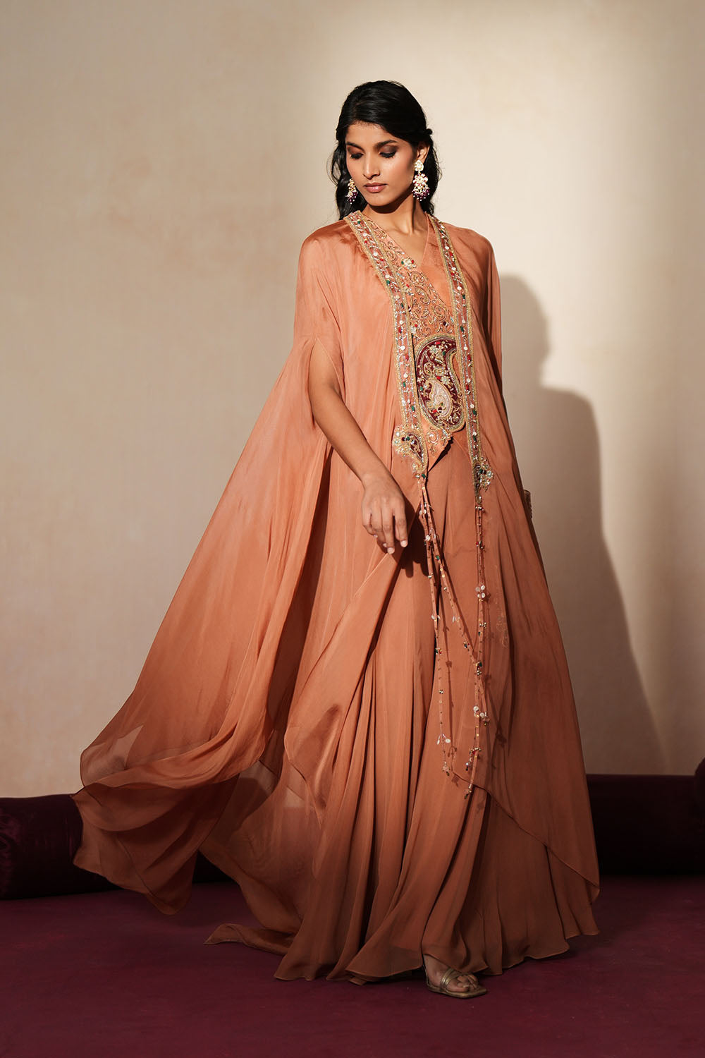 Woman in a metallic mocha  traditional outfit standing againsta beige wall on a red carpet.