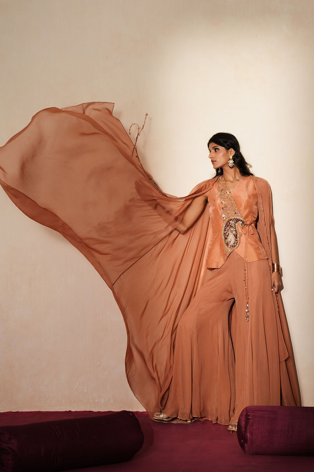 Woman in a metallic mocha  traditional outfit standing againsta beige wall on a red carpet.