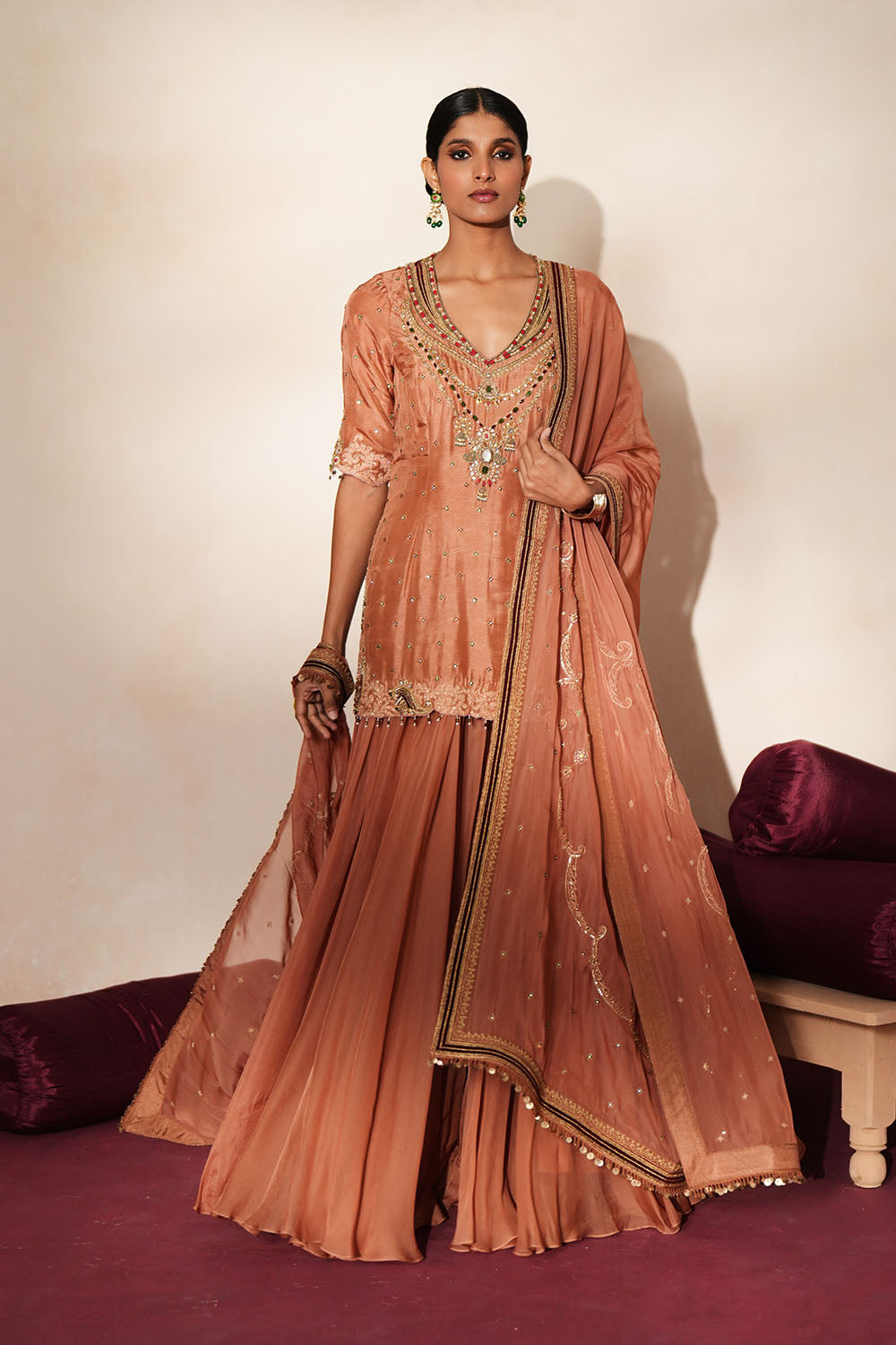 Woman in a metallic mocha  traditional outfit standing againsta beige wall on a red carpet.