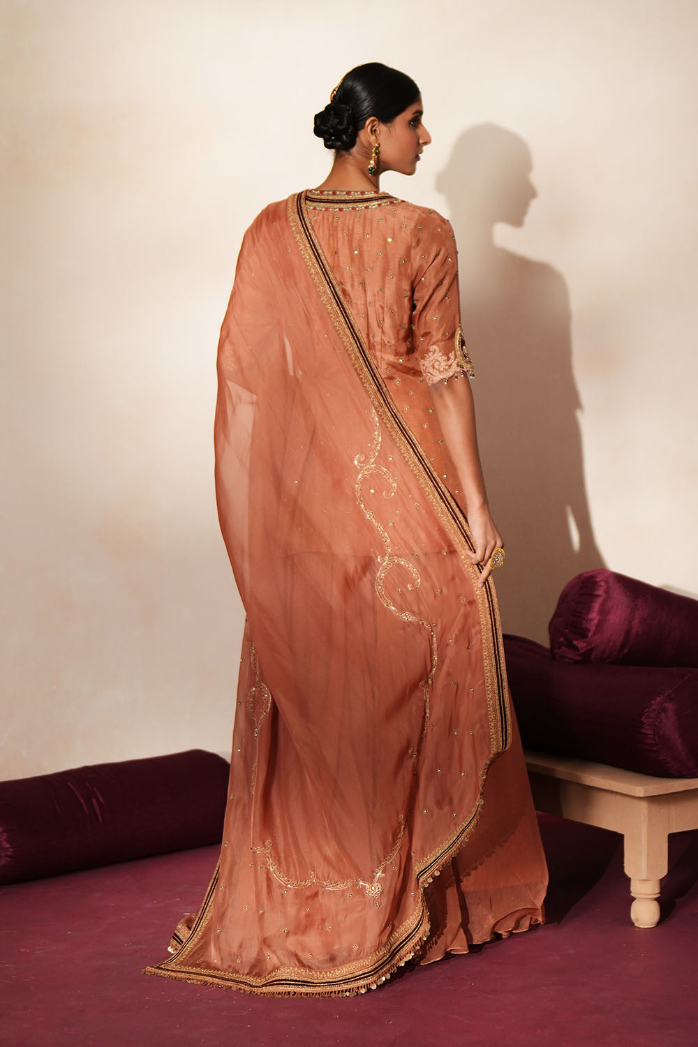 Woman in a metallic mocha  traditional outfit standing againsta beige wall on a red carpet.
