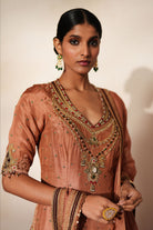 Woman in a metallic mocha  traditional outfit standing againsta beige wall on a red carpet.