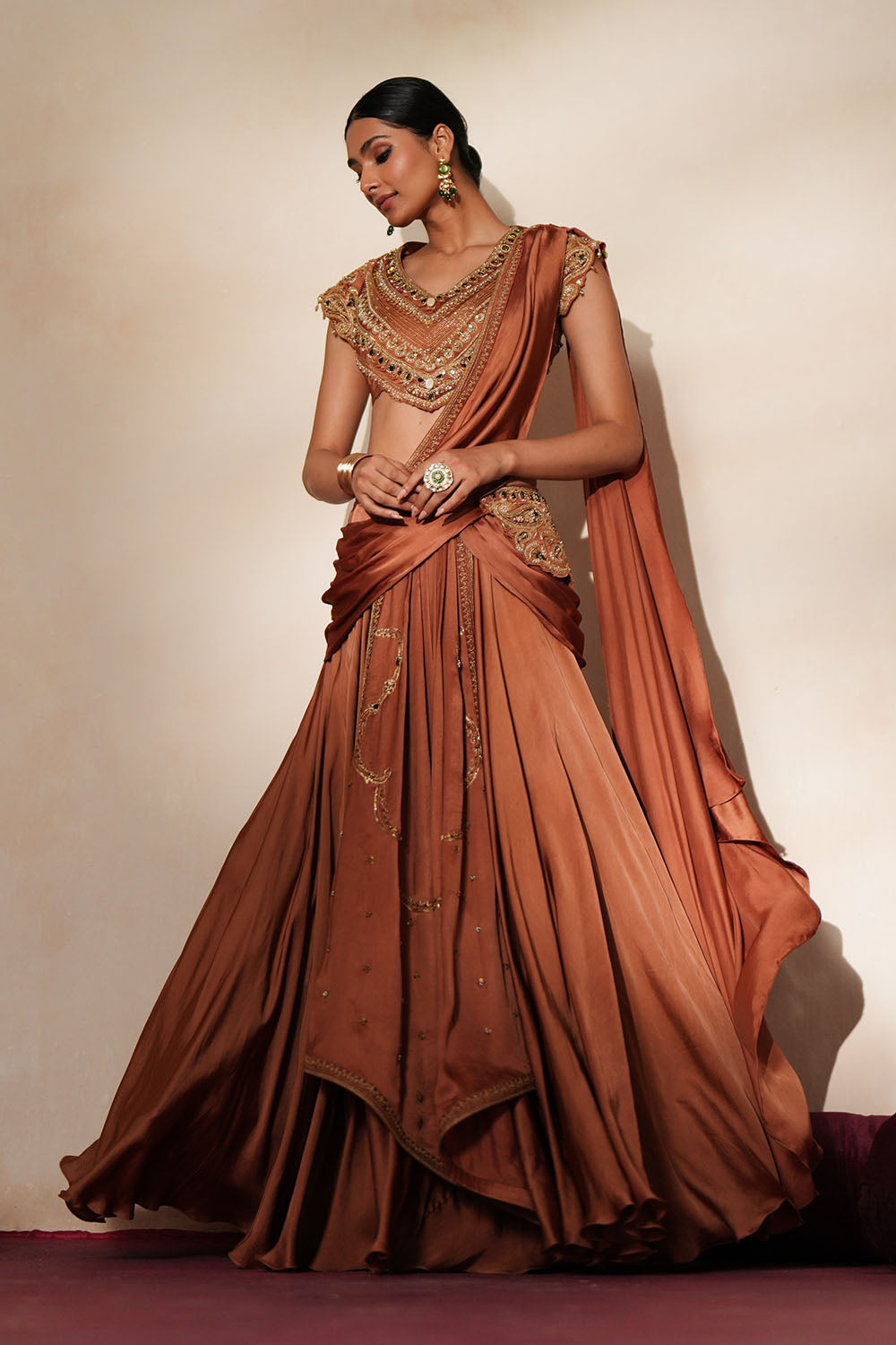 Woman in a metallic mocha  traditional outfit standing againsta beige wall on a red carpet.