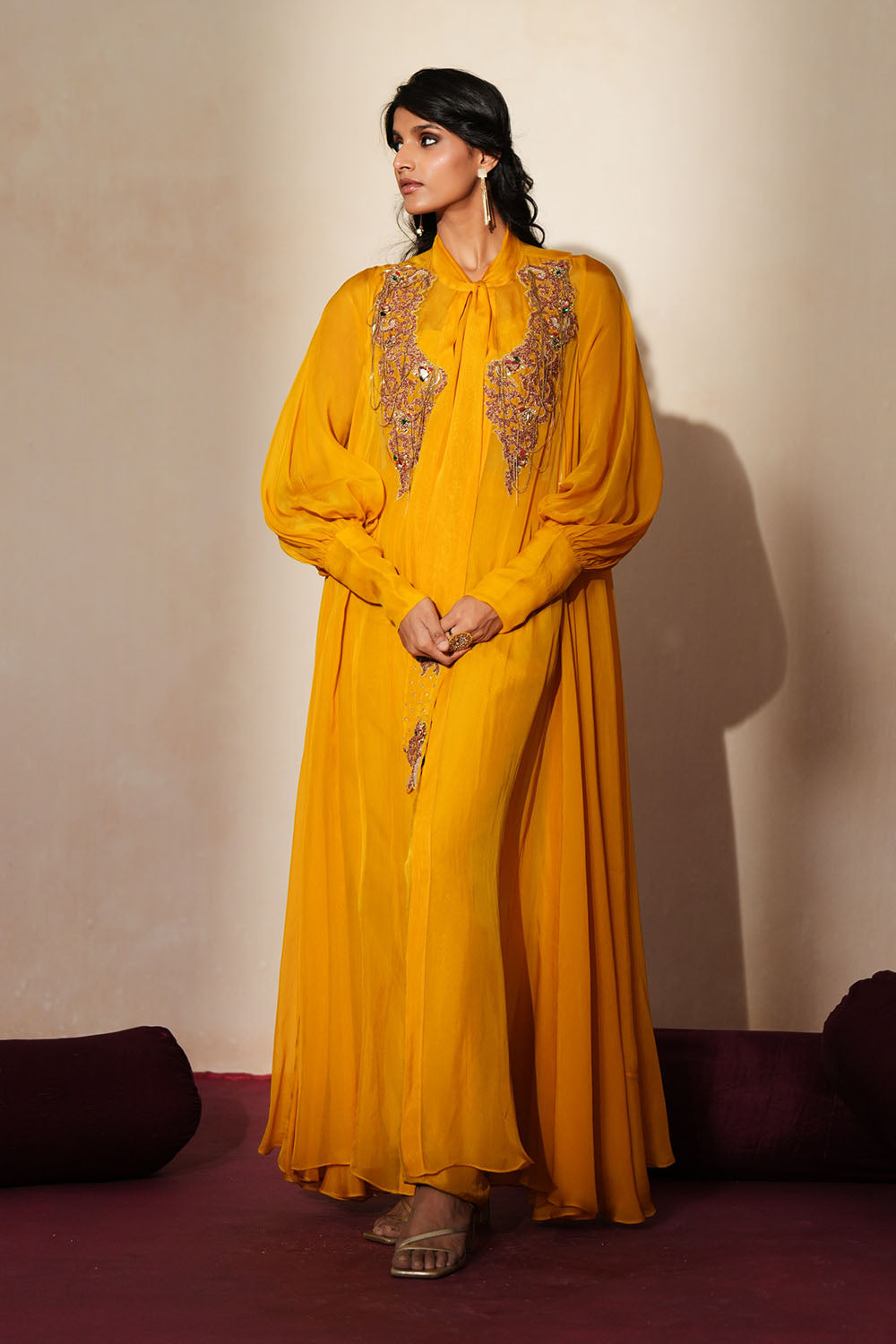 Woman in a golden yellow traditional outfit standing againsta beige wall on a red carpet.