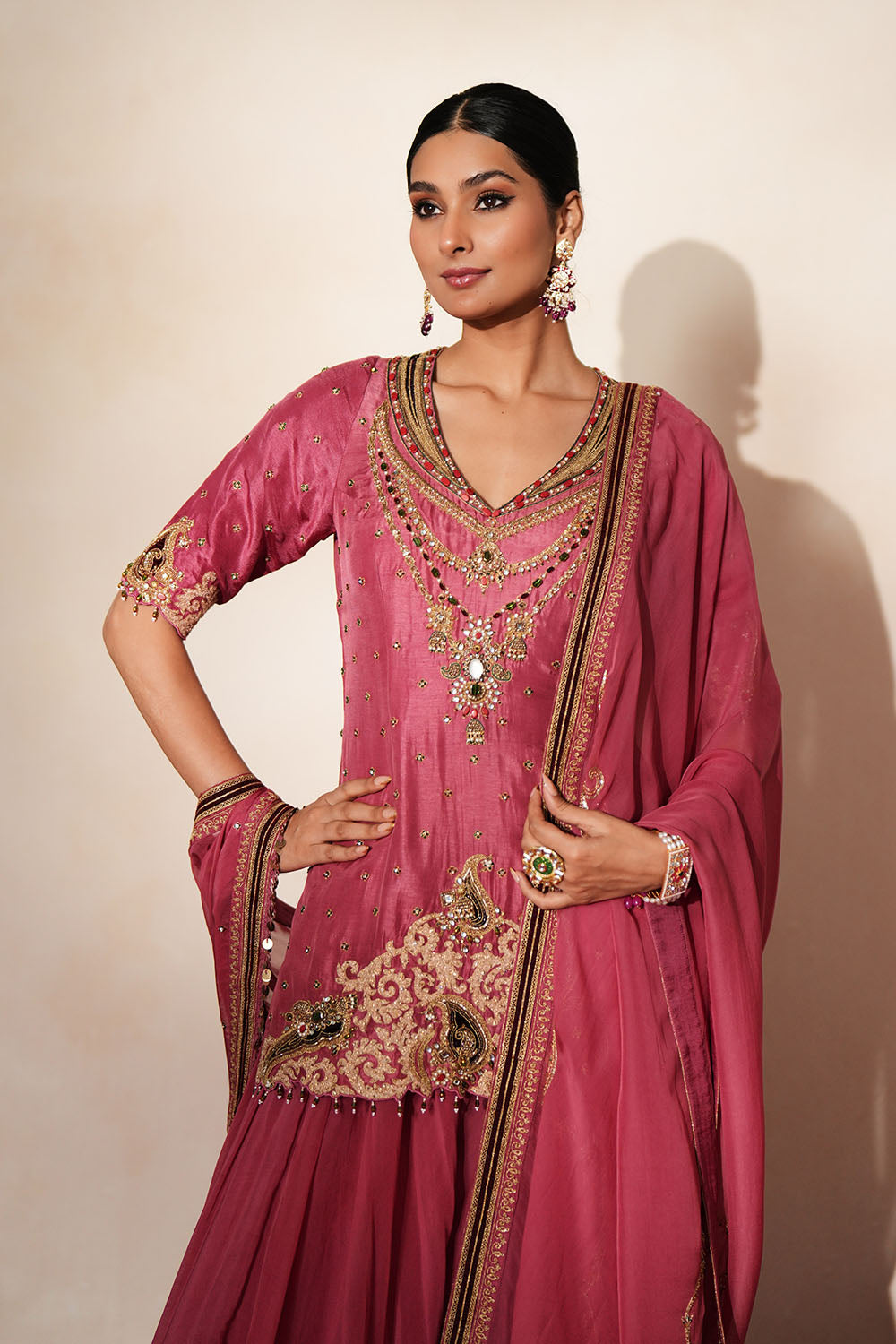 Woman in a mauve pink traditional outfit standing againsta beige wall on a red carpet.
